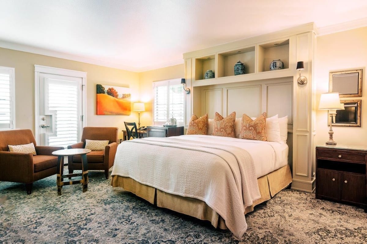 A hotel room with a large bed, a chair, a small table, a desk, and a lamp.