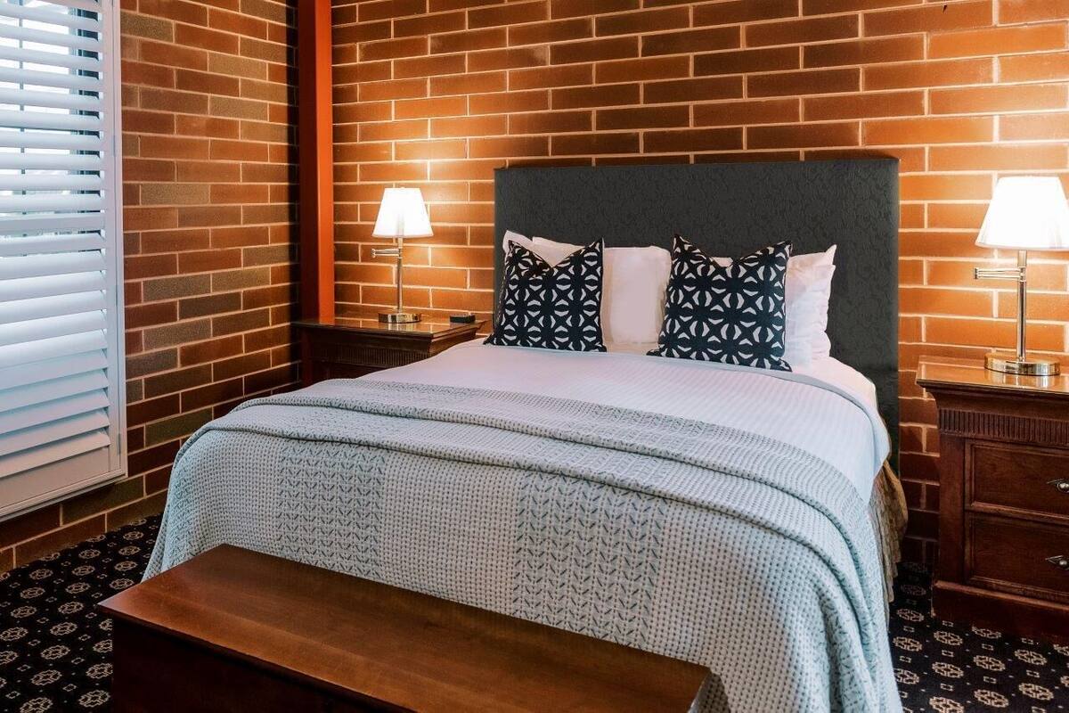 A hotel room with a brick wall, a bed with patterned pillows, bedside lamps, and a wooden bench.