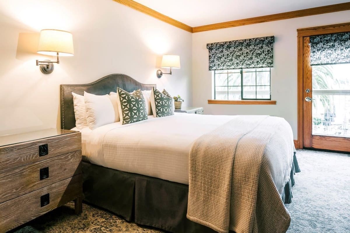 A bedroom with a bed, bedside lamps, a window with a patterned valance, and a wooden door.