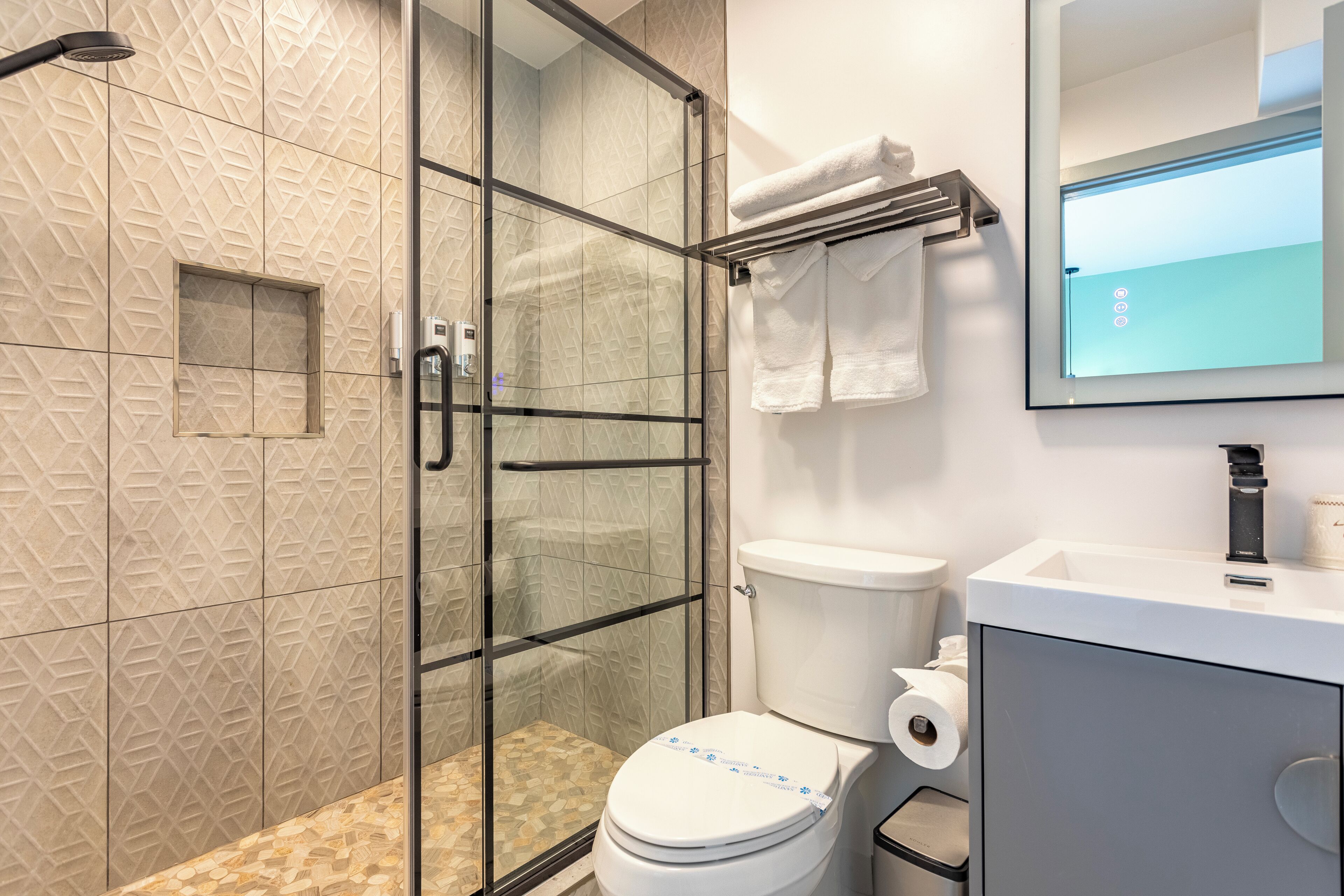 Un baño moderno con ducha envuelta en vidrio, un inodoro con soporte para toallas de papel y un lavamanos con espejo arriba.