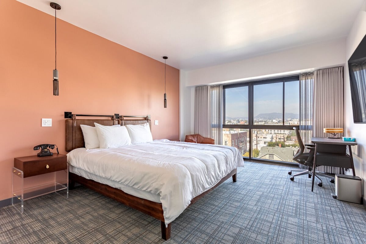 A hotel room with a large bed, a desk, and a view of the city through large windows.
