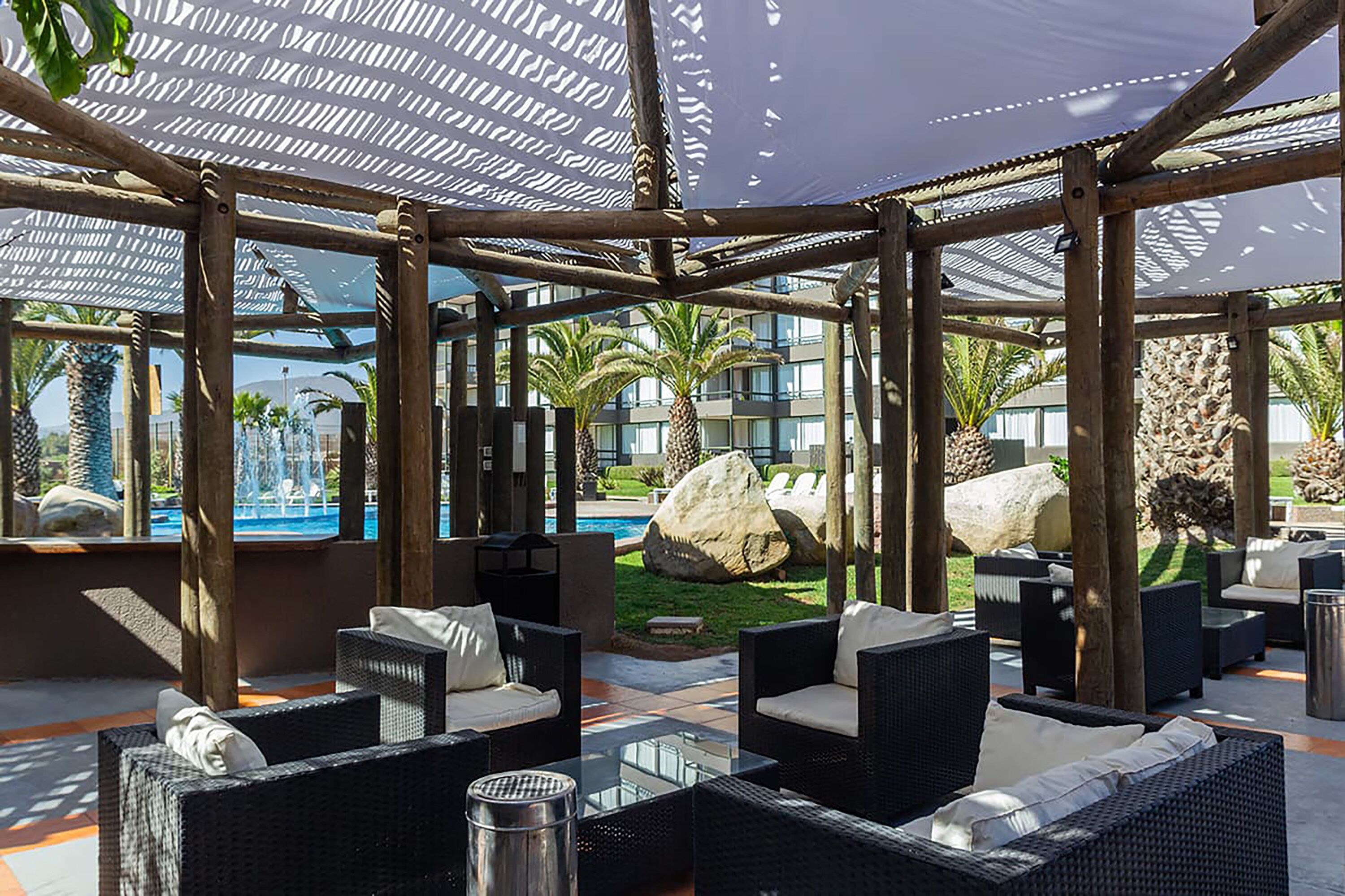 A covered outdoor seating area with wicker furniture and a view of a pool and palm trees.