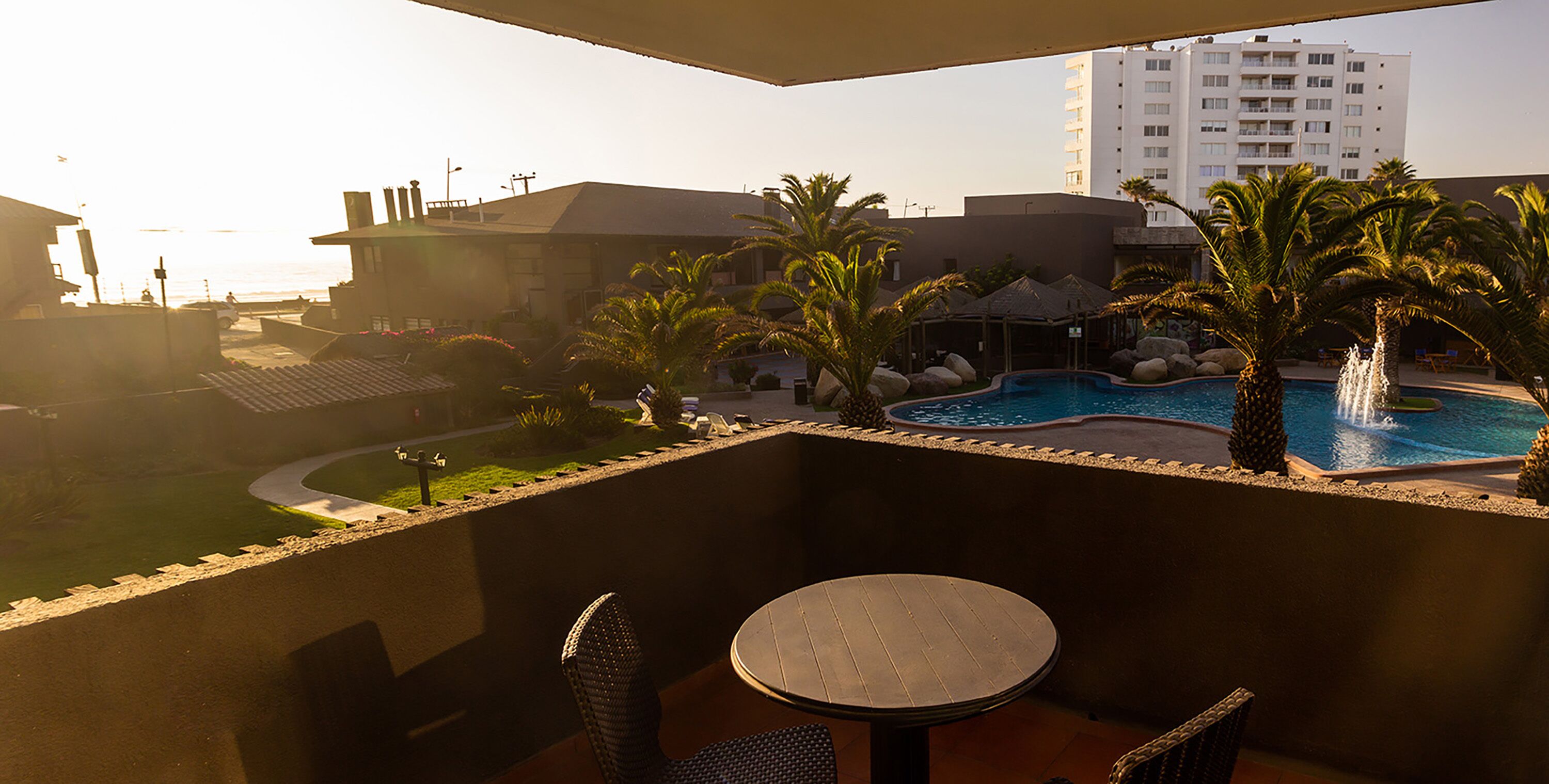 A balcony with a pool, palm trees, and a view of a hotel building.
