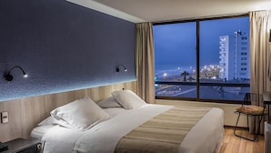 Habitación de hotel con una cama grande, una mesita de noche, una silla y vista a la ciudad al atardecer.