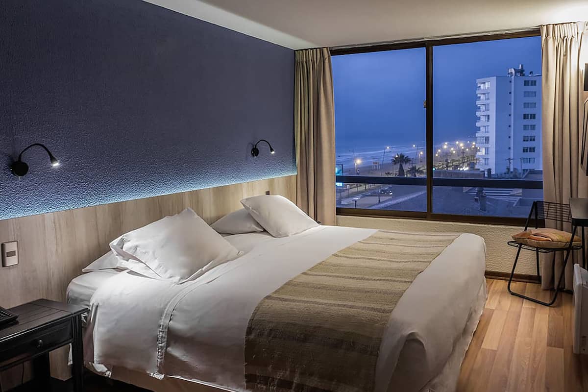 Une chambre d’hôtel avec un grand lit, une table de chevet, une chaise et une vue sur la ville au crépuscule.