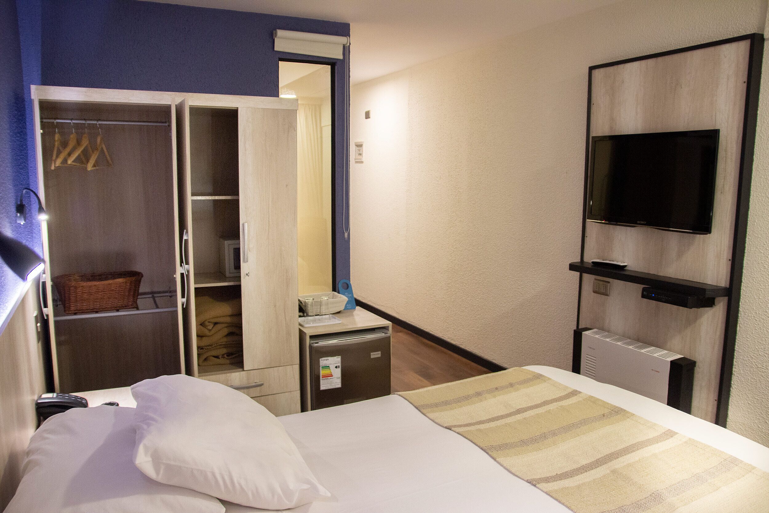 Een hotelkamer met een bed, een kledingkast, een bureau met lamp, een televisie en een kleine koelkast.