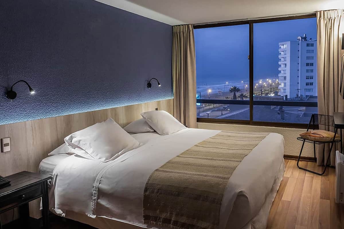 Une chambre d’hôtel avec un grand lit, une table de chevet, un bureau, une chaise et une grande fenêtre donnant sur la ville.