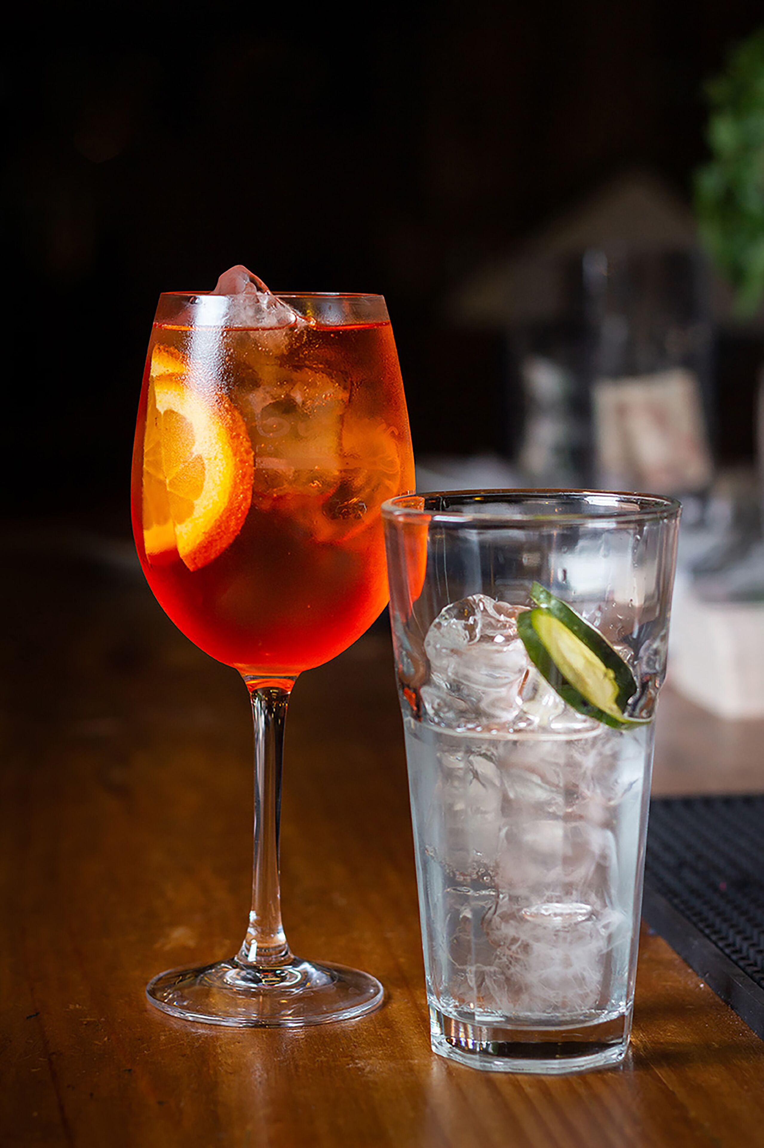 Two glasses of beverages, one with ice and a slice of orange, and the other with ice and a slice of lime, on a wooden table.