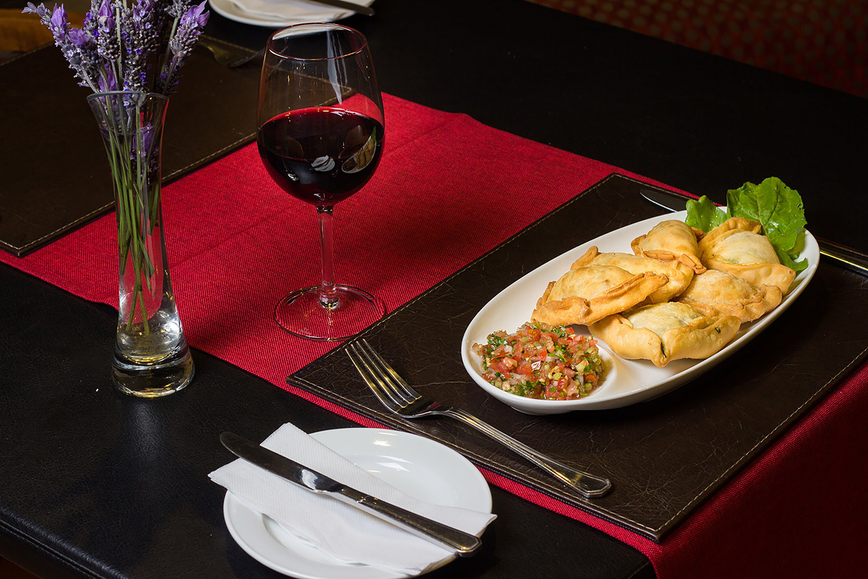 A table set with a red placemat, a white plate with pastries, a glass of red wine, and a vase with flowers.