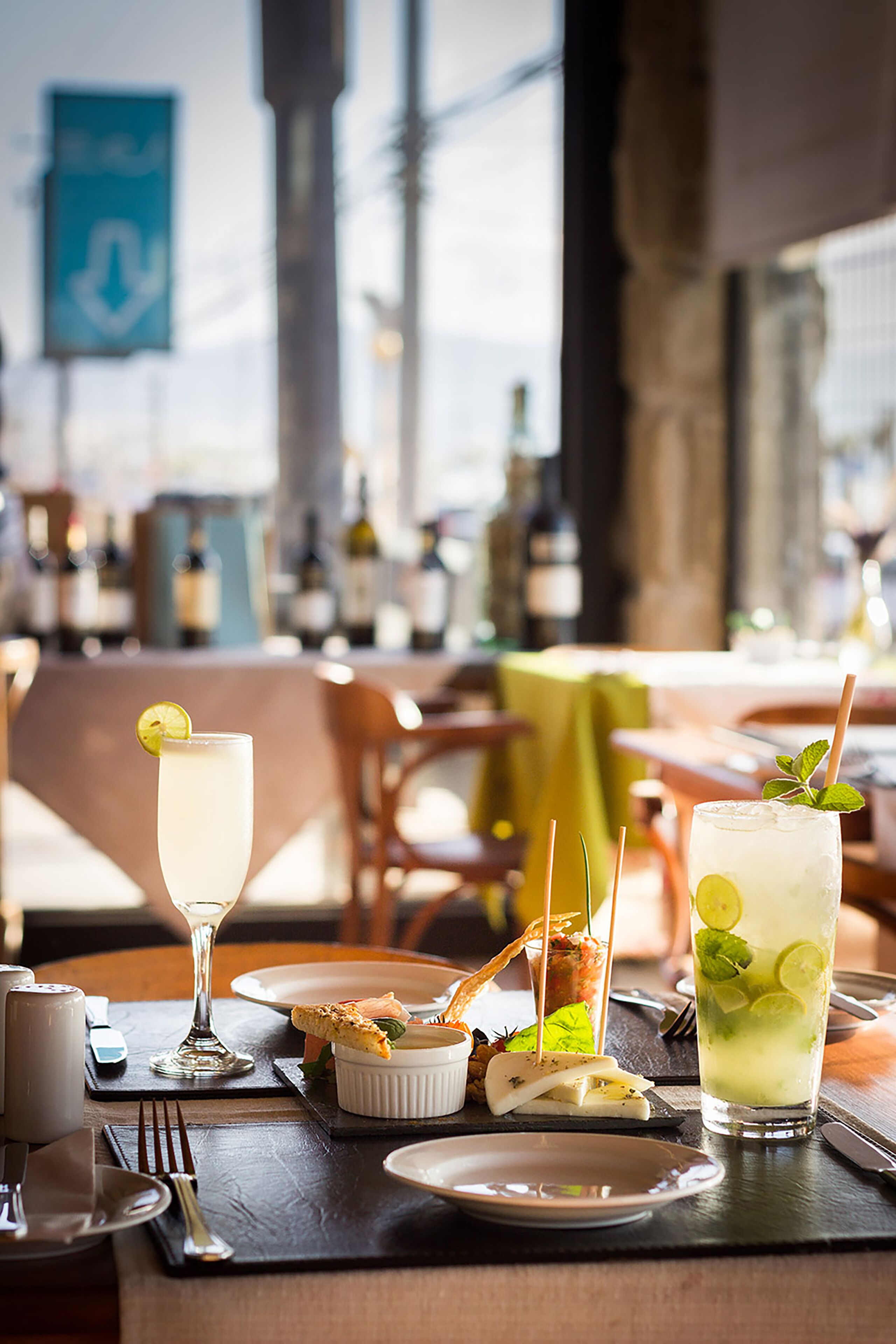 Uma mesa de restaurante com um copo de vinho branco, um cocktail com limão e hortelã, e um pequeno prato de queijo.