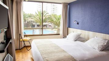 Une chambre d’hôtel avec un grand lit, une télévision, un fauteuil et une vue sur une piscine et des palmiers.