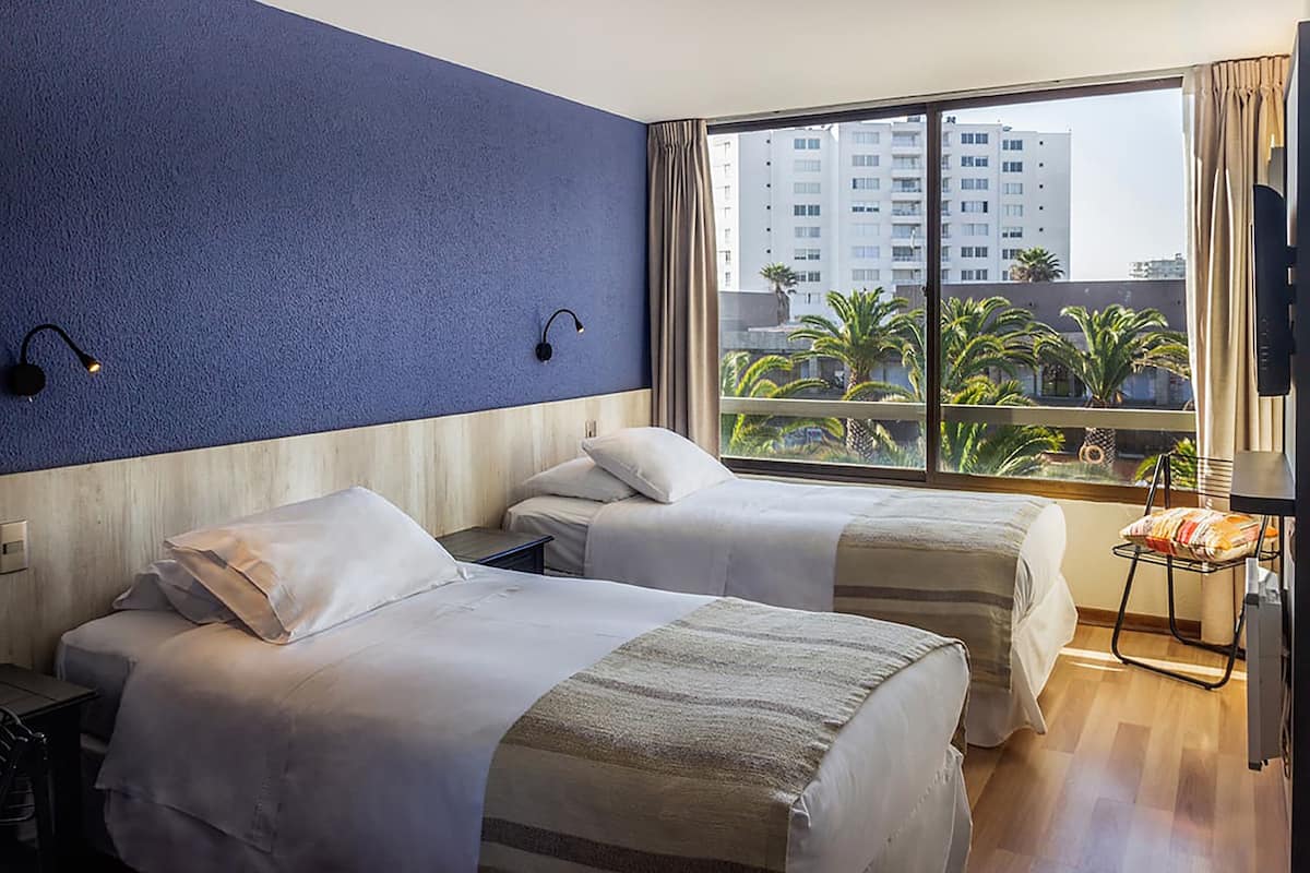 Une chambre d’hôtel avec deux lits, une grande fenêtre donnant sur des palmiers et des immeubles, et une petite table avec une chaise.