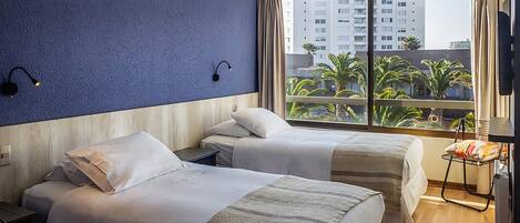 A hotel room with two beds, a large window with a view of palm trees and buildings, and a small table with a chair.