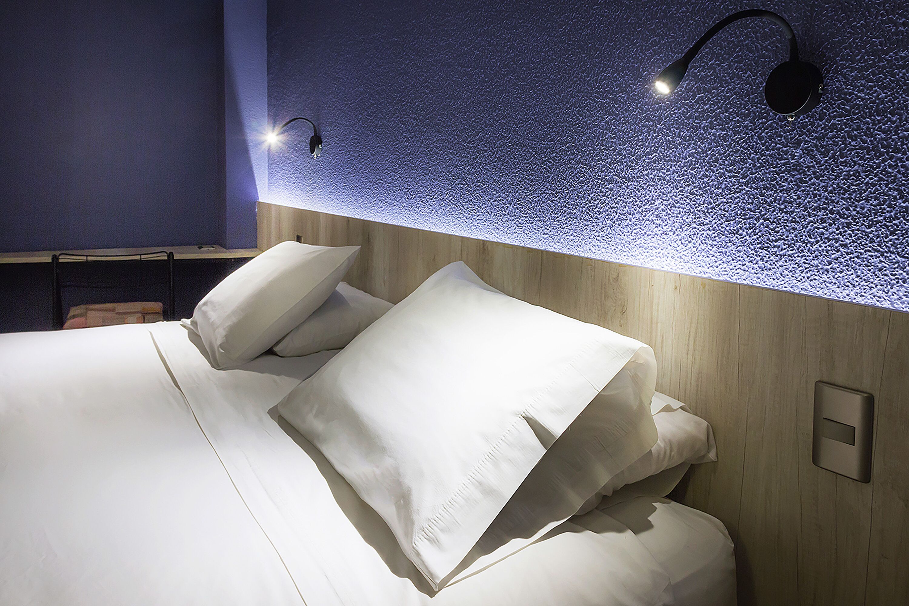 A neatly made bed with white linens and pillows, a wooden headboard, and two wall-mounted reading lights.