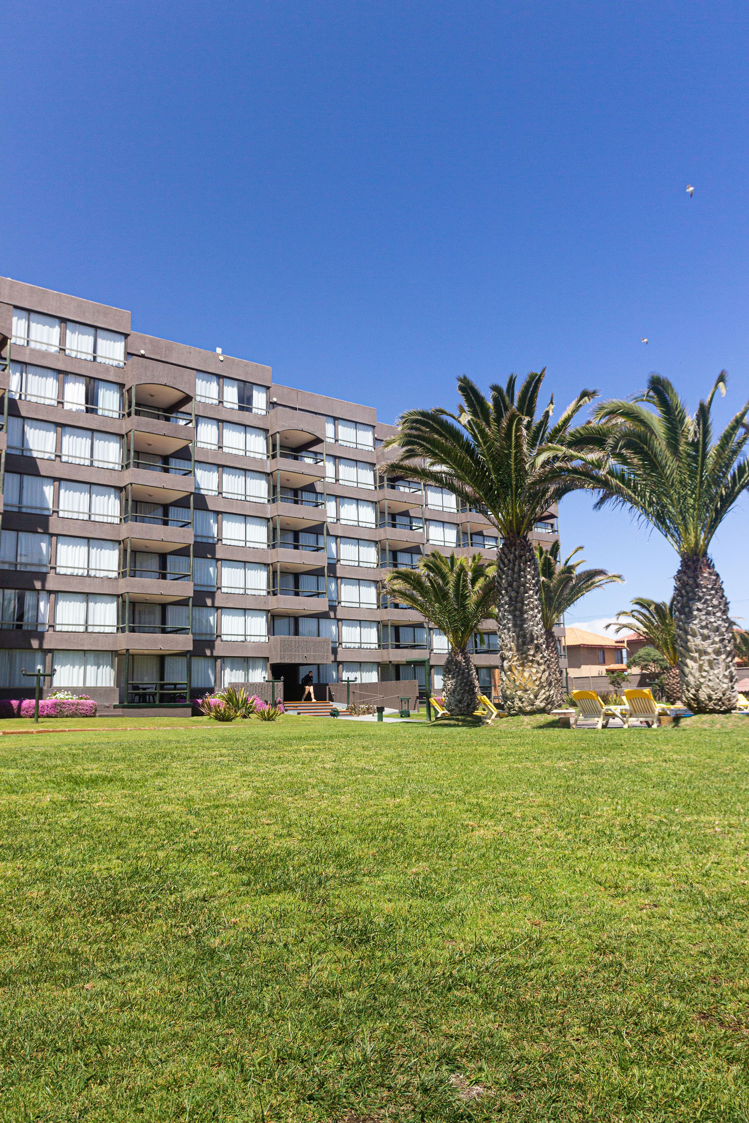A multi-story hotel with balconies, a grassy lawn, and palm trees.