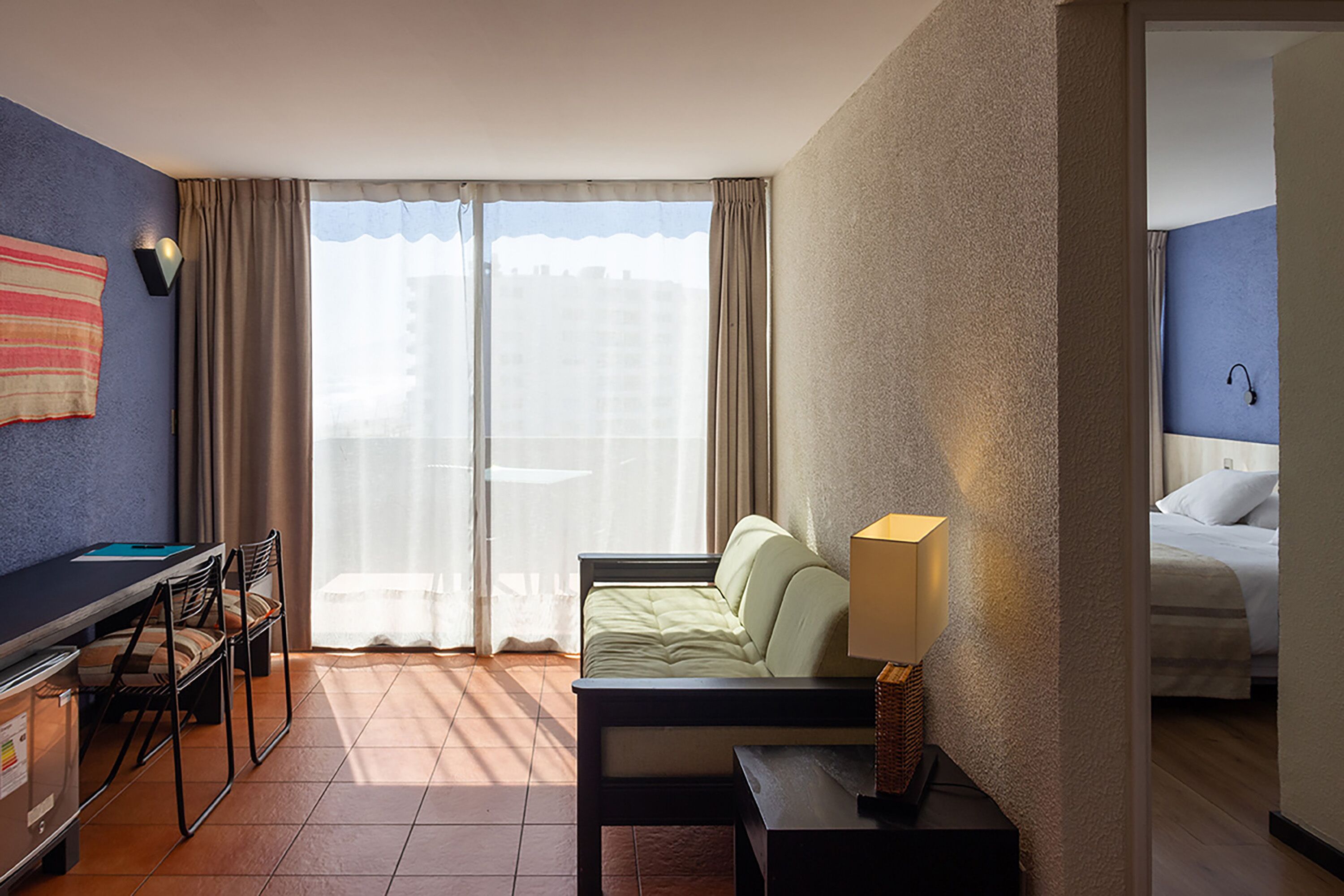 A hotel room with a bed, a desk with a chair, a lamp, and a view through the window.