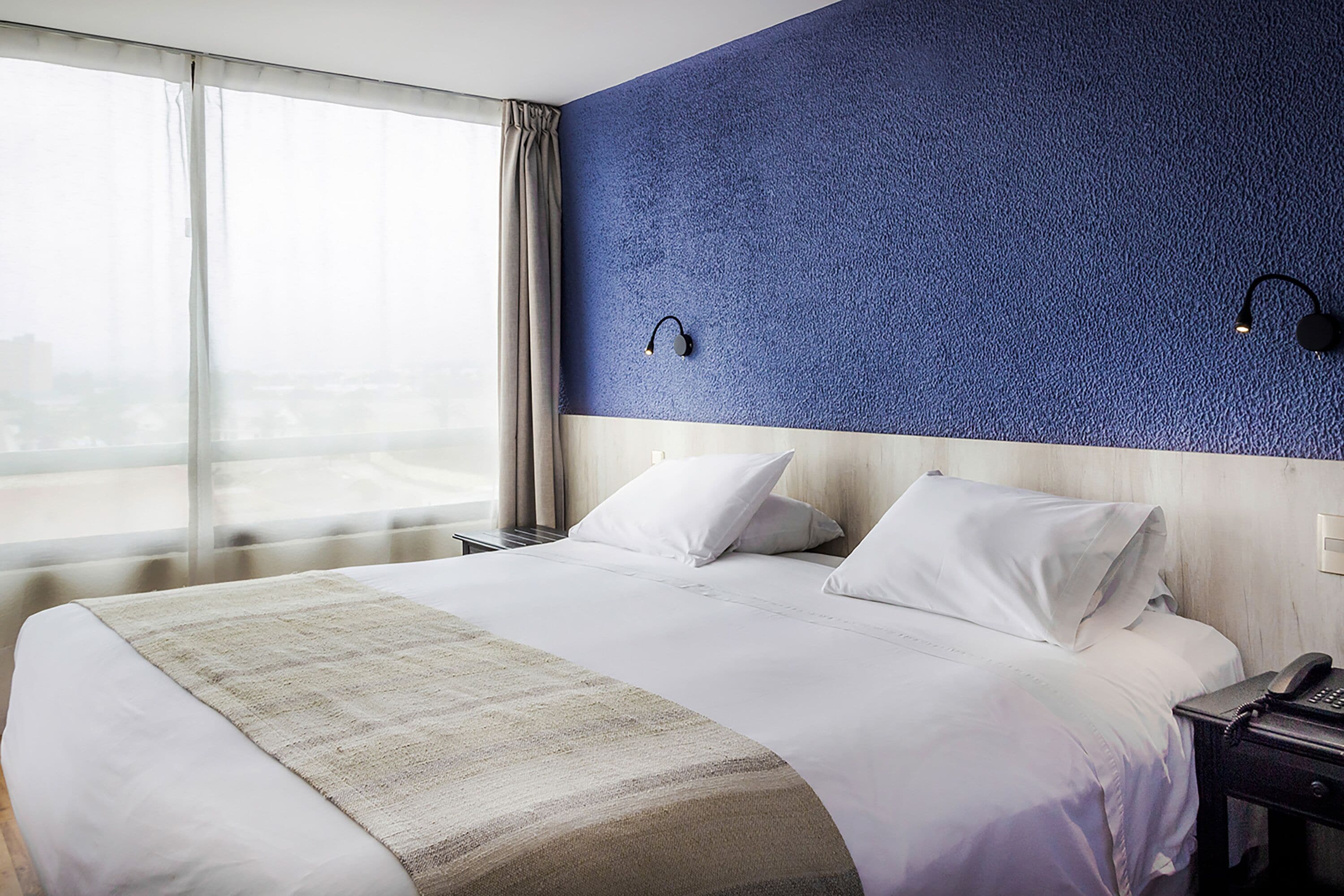Une chambre d’hôtel avec un grand lit, une table de chevet, un téléphone et une fenêtre avec des rideaux.