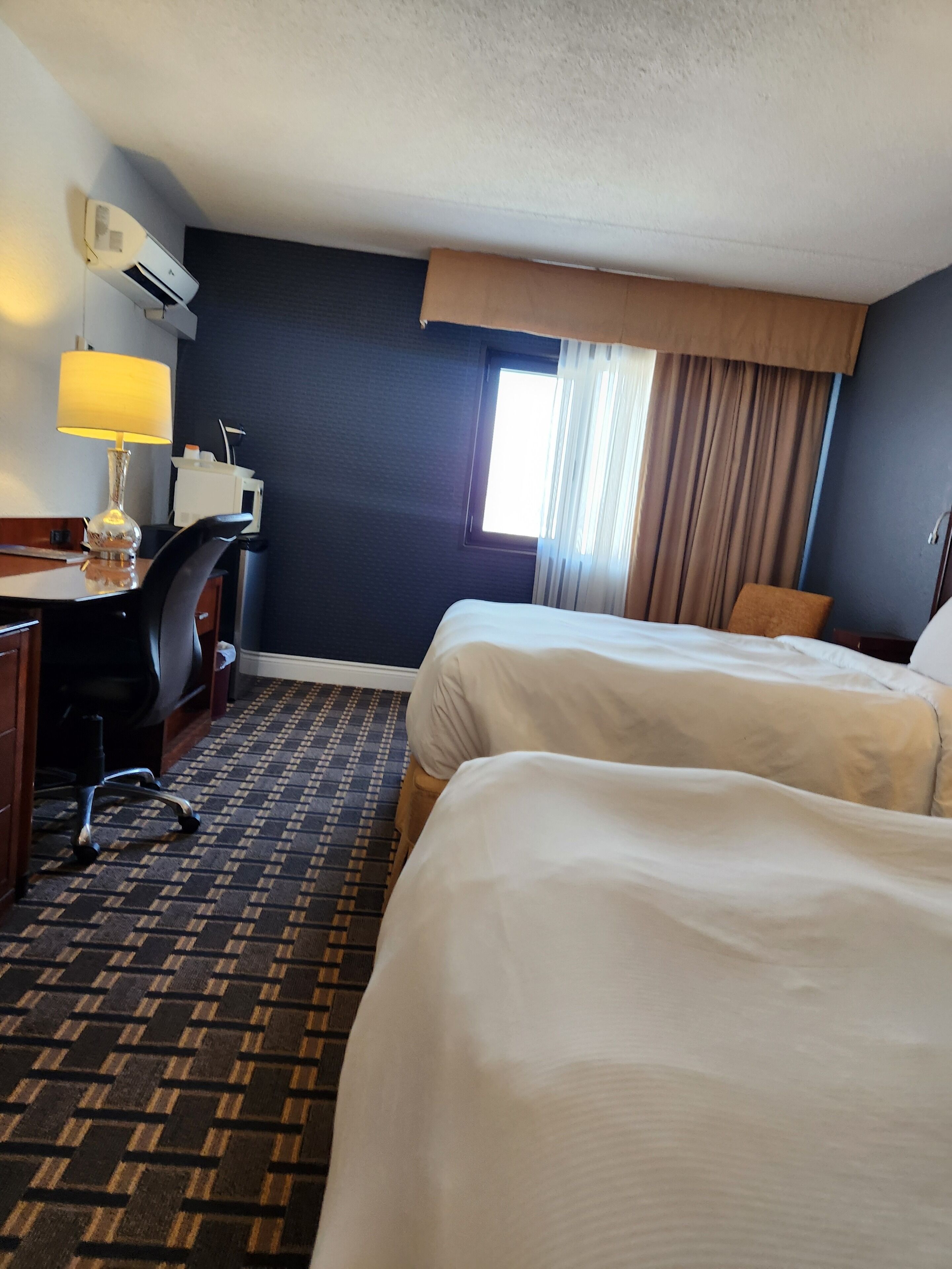 A hotel room with two beds, a desk with a lamp, an air conditioner, and a window with curtains.