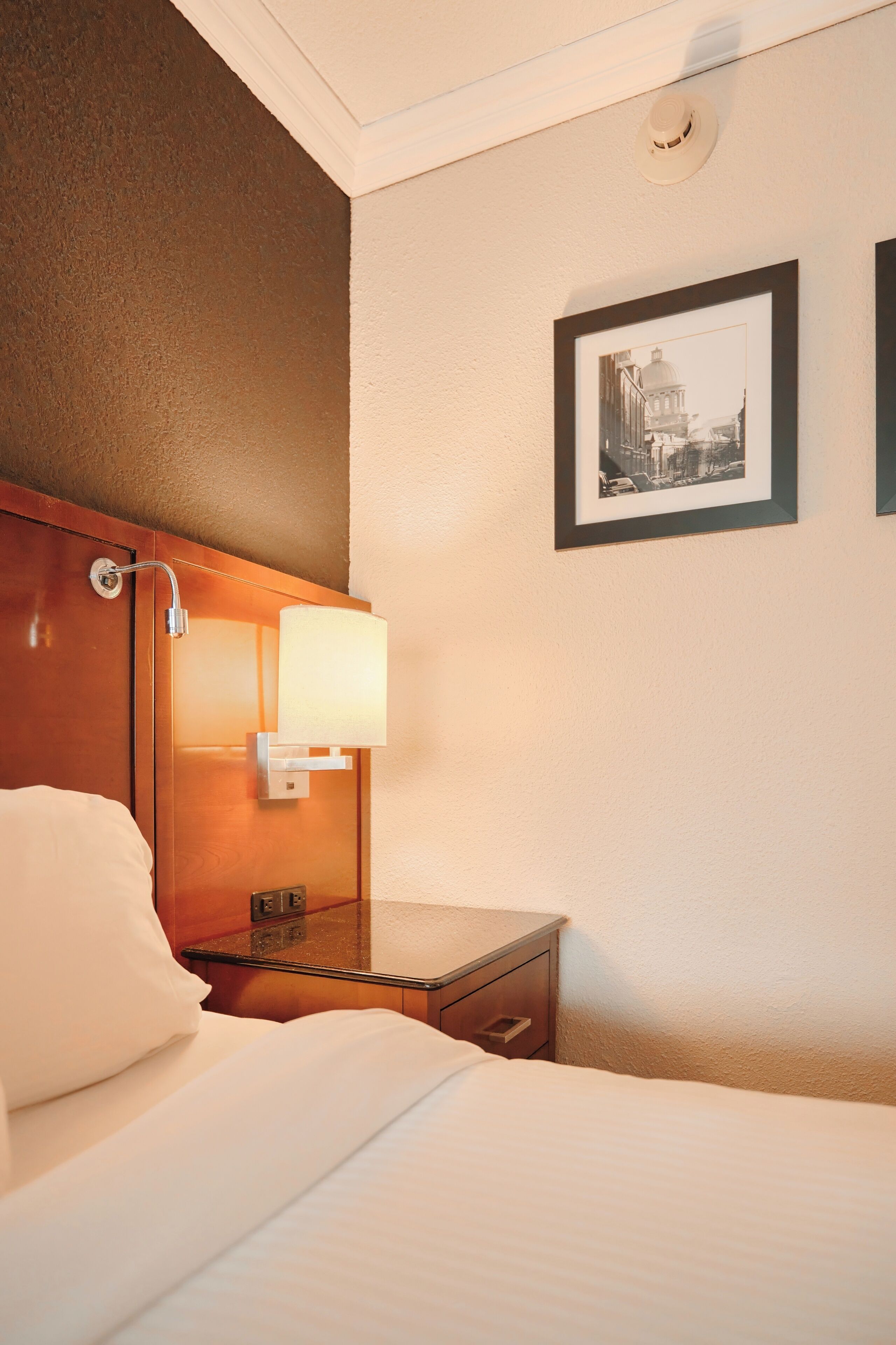 A hotel room with a bed, a nightstand, a wall-mounted lamp, and a framed picture on the wall.