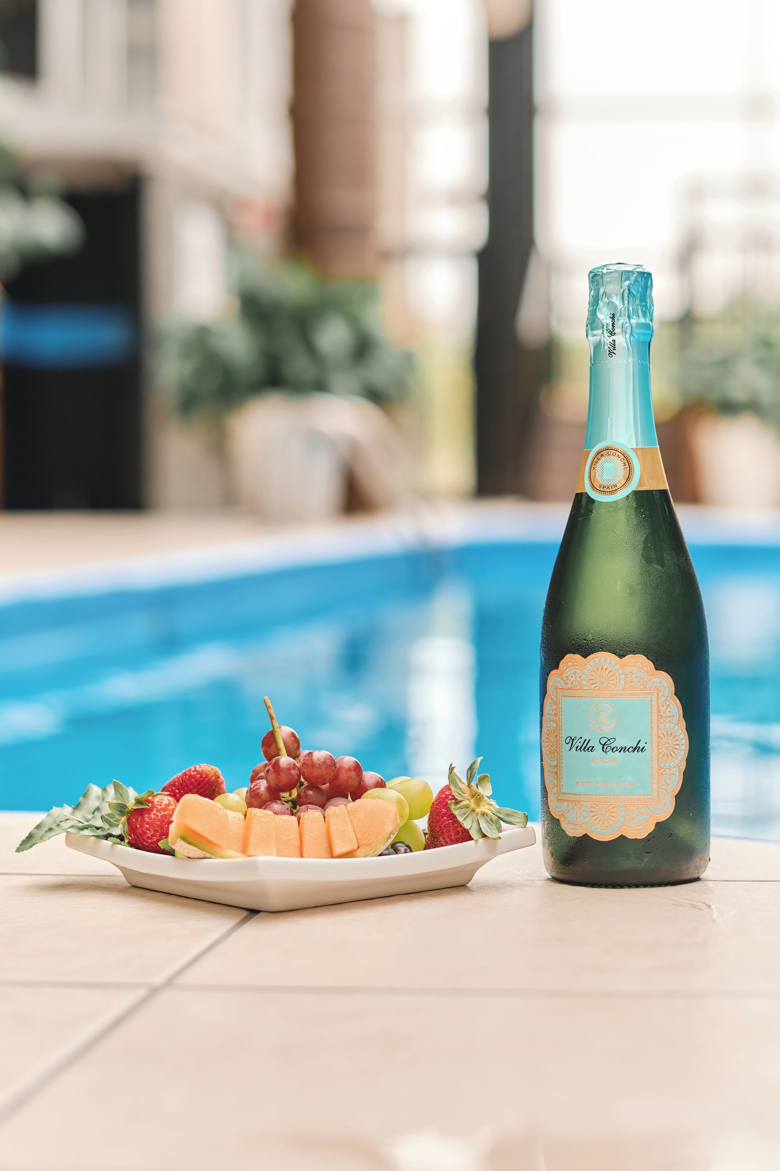 A bottle of Villa Conchi champagne next to a plate of fruit by a pool.