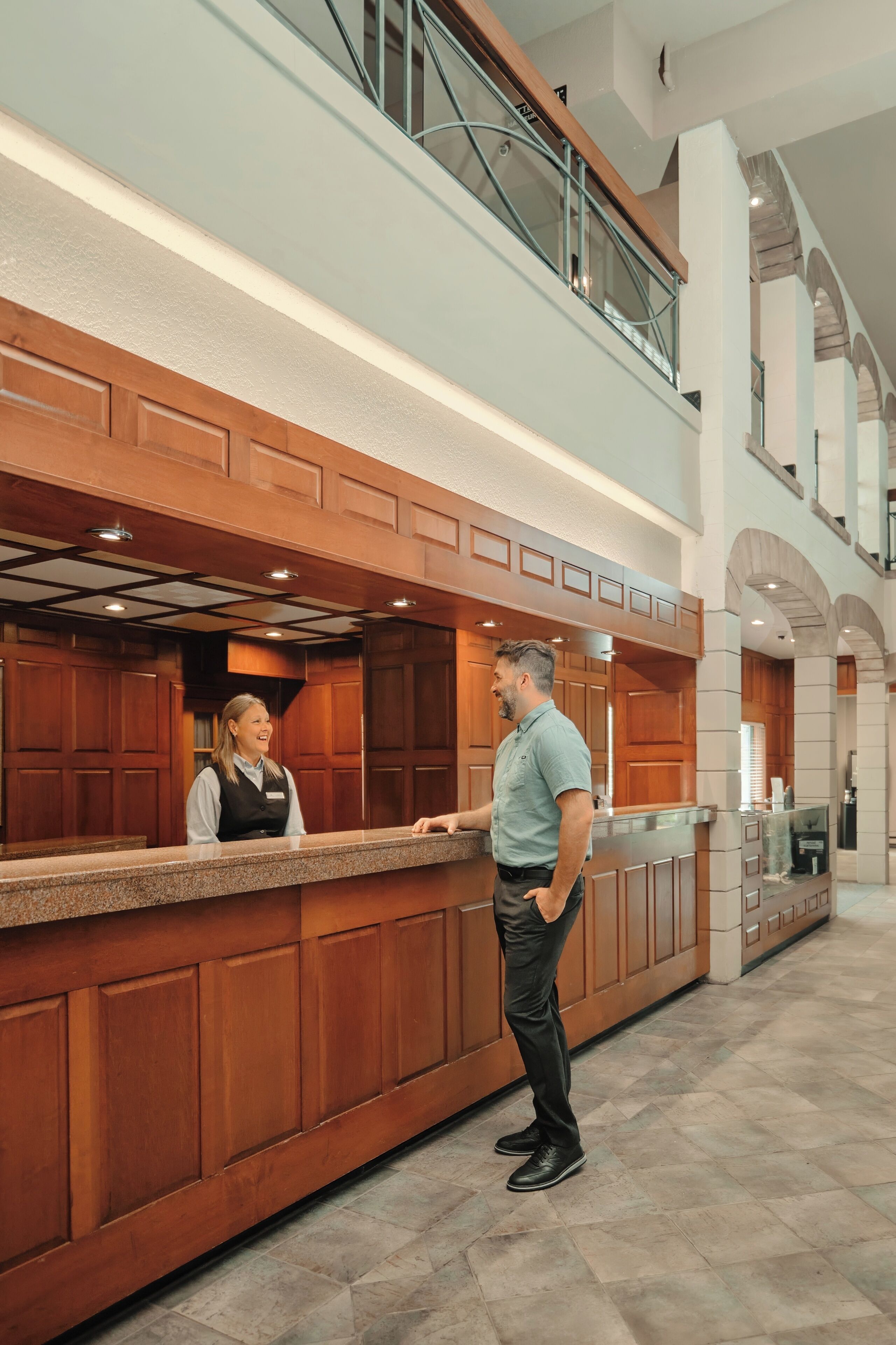 A hotel reception area with a double-height wooden counter, a glass railing, and a high ceiling with arched windows.