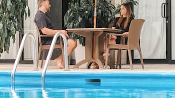 Zona junto a la piscina con mesa y sillas bajo una sombrilla, rodeada de plantas y la fachada de un edificio.