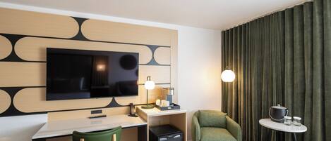 Habitación de hotel con televisión de pantalla plana montada en la pared, escritorio con lámpara, refrigerador pequeño, una silla verde y una mesita de noche con hervidor y tazas.