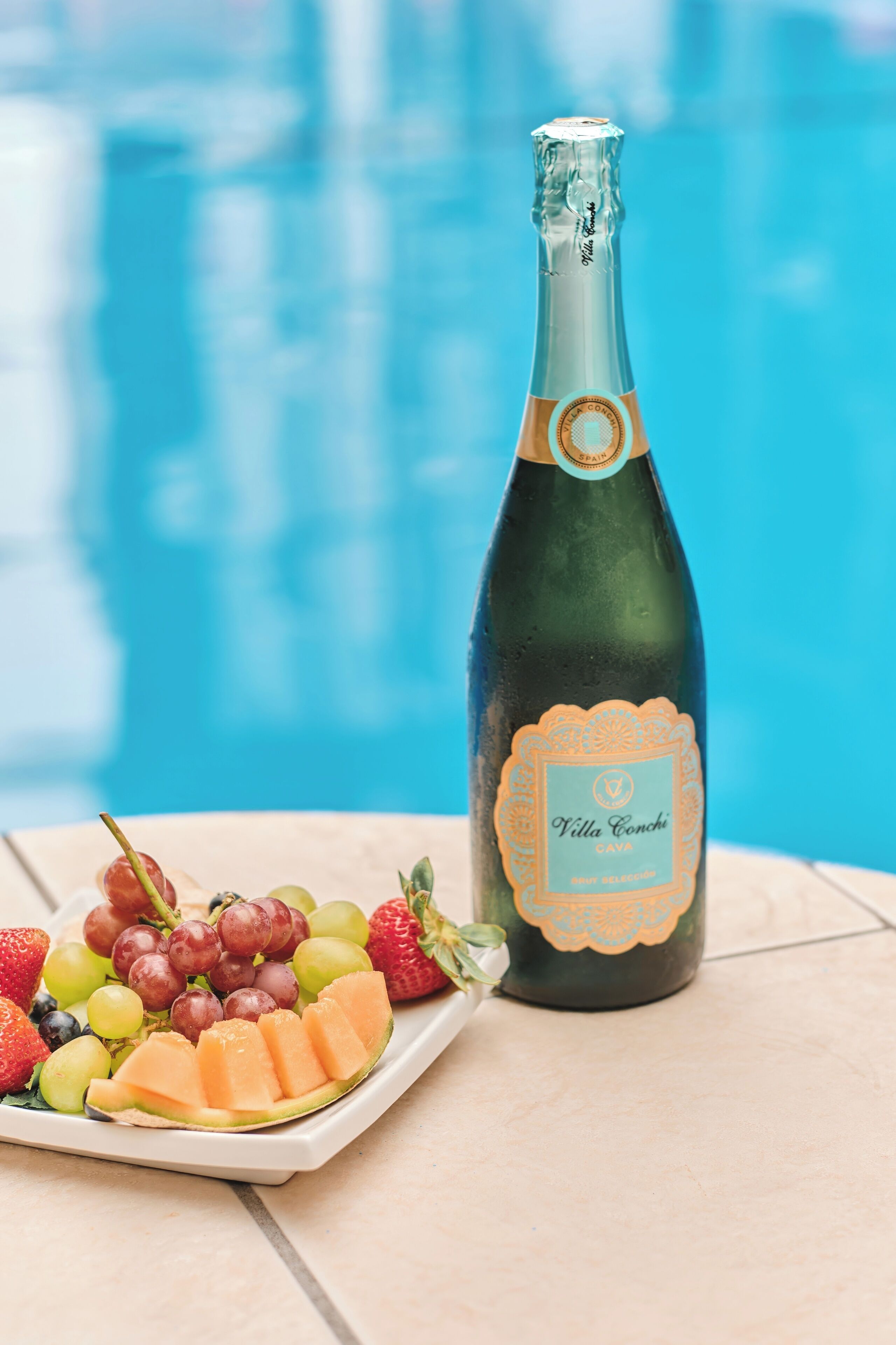 A bottle of Villa Conchi Cava wine and a fruit platter by a poolside.