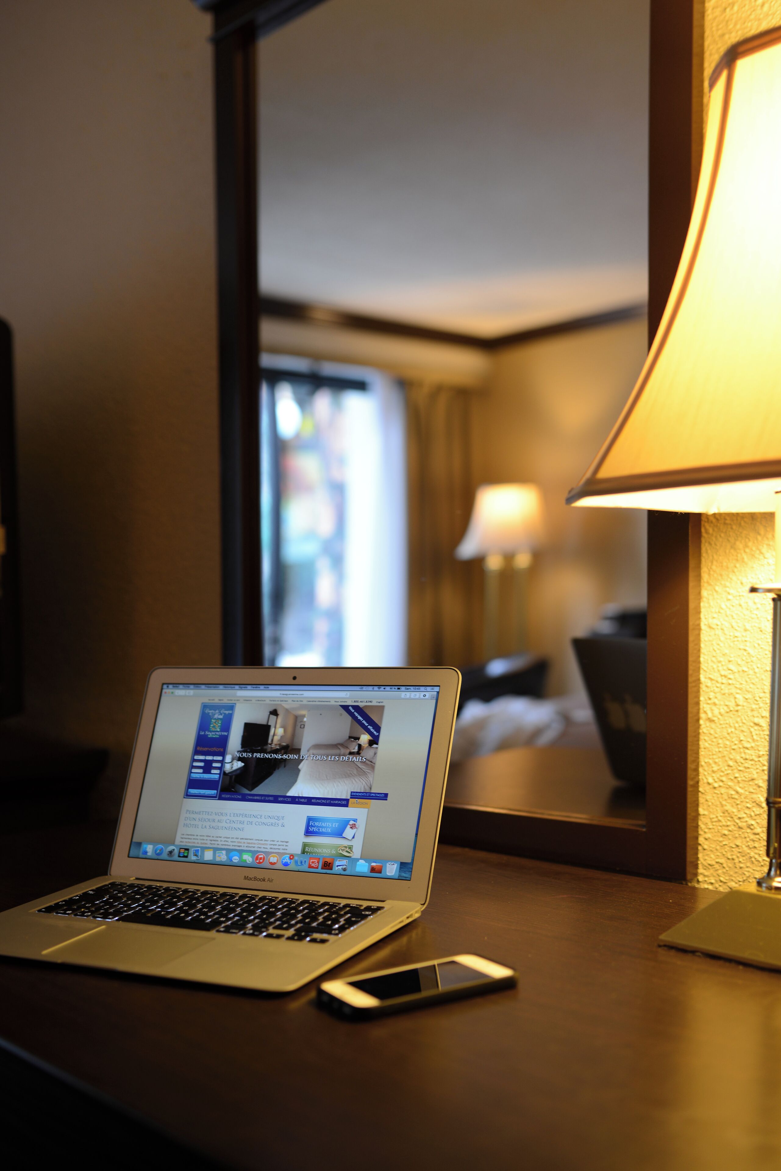 A hotel room with a laptop on a desk, a bedside lamp, and a bed with a headboard.