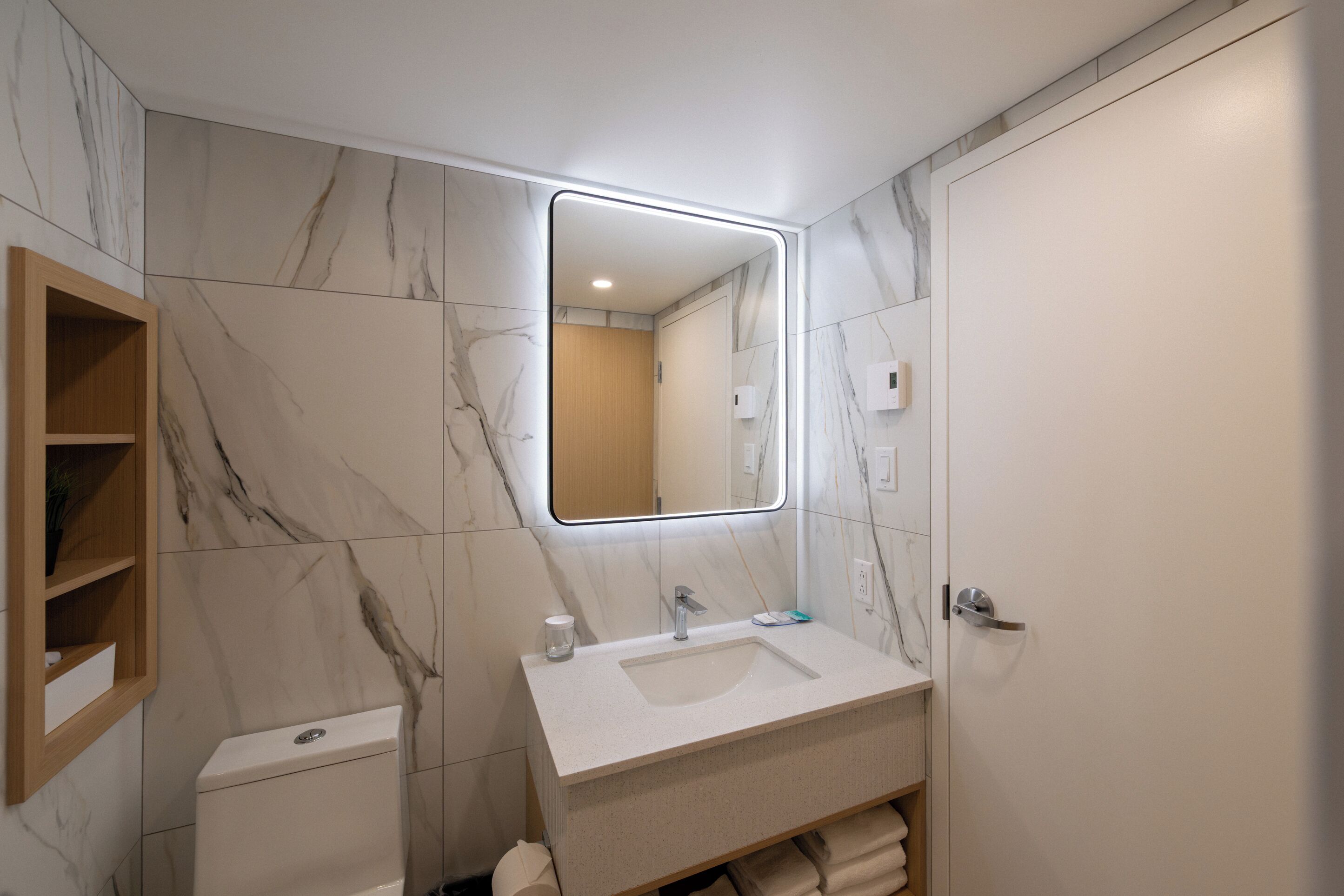 Un baño moderno con pared de mármol, estante de madera, inodoro, lavabo con espejo y perchero.