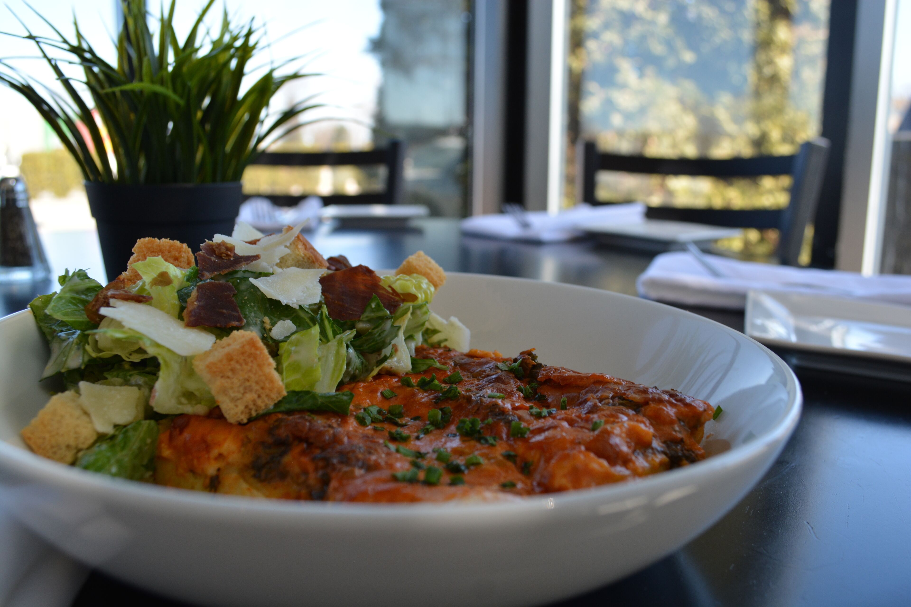 A bowl of salad with croutons and a tomato-based sauce, placed on a table with a potted plant in the background.