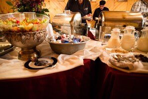 A buffet table with a variety of food items, including a large bowl of fruit salad, a metal container with snacks, and several pitchers of beverages. - Alta Peruvian Lodge (Sandy)