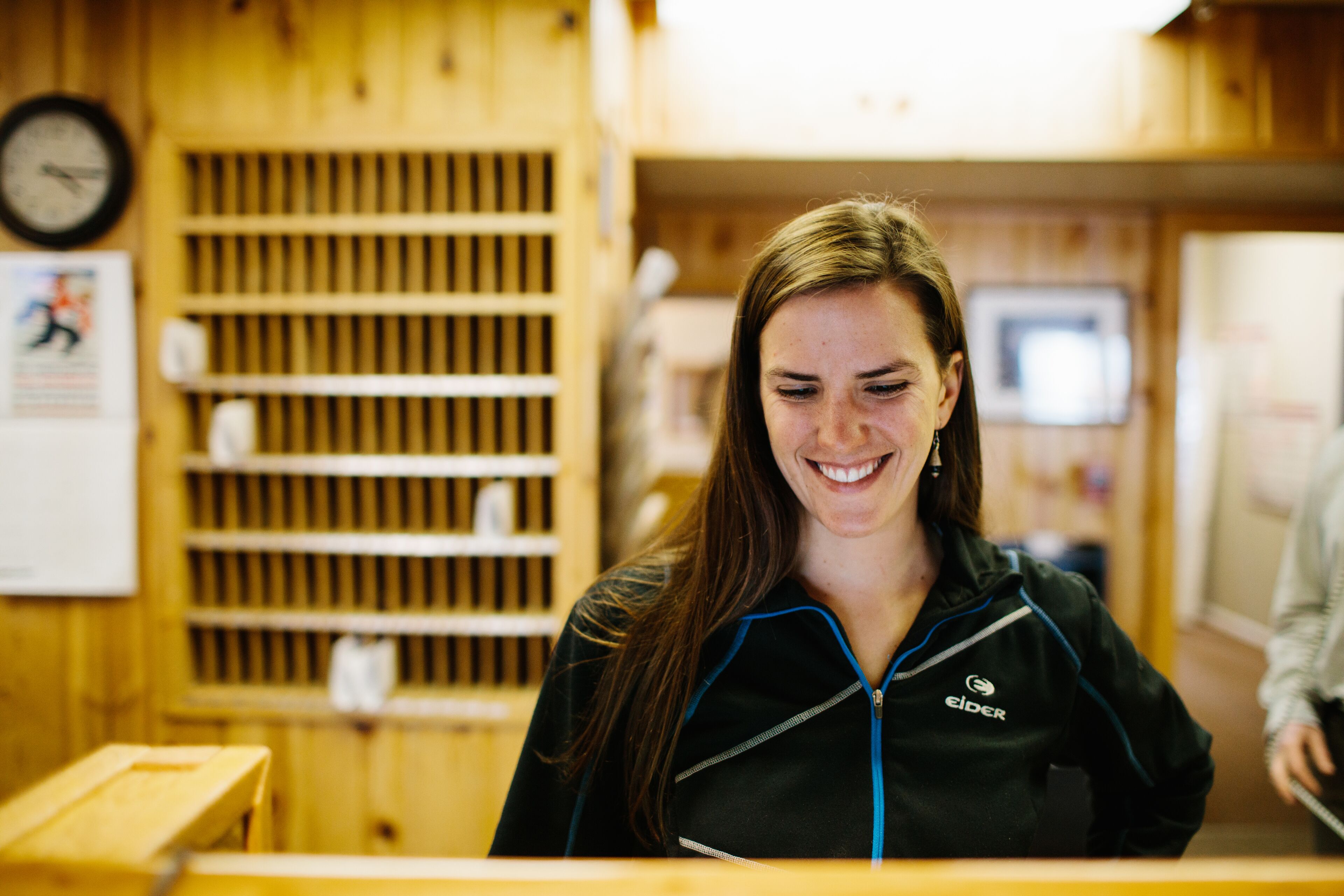 A woman in a black jacket with a zipper and logo, standing in a room with wooden walls and shelves.