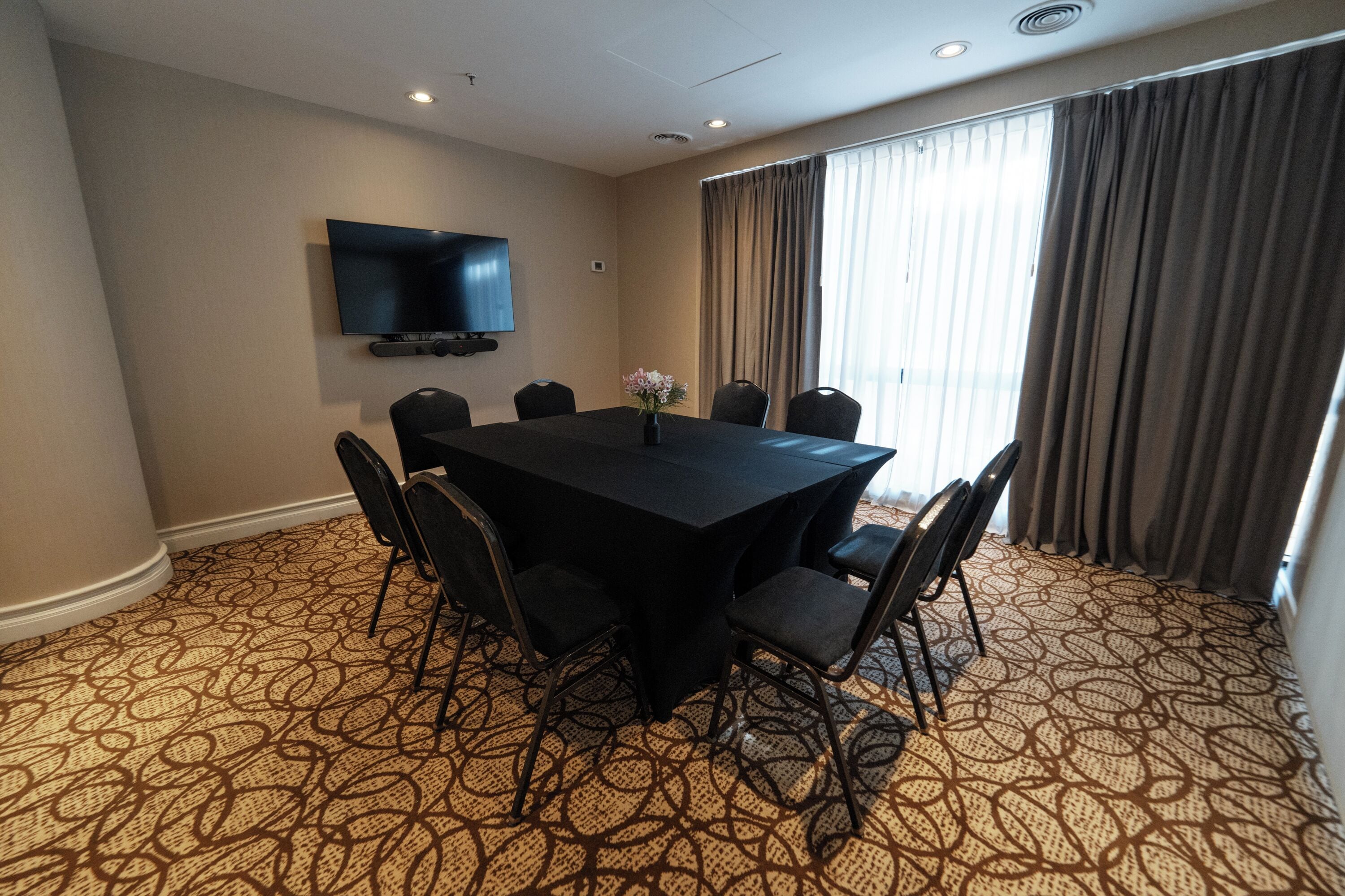 A conference room with a large table, six chairs, a flat-screen TV mounted on the wall, and a patterned carpet.