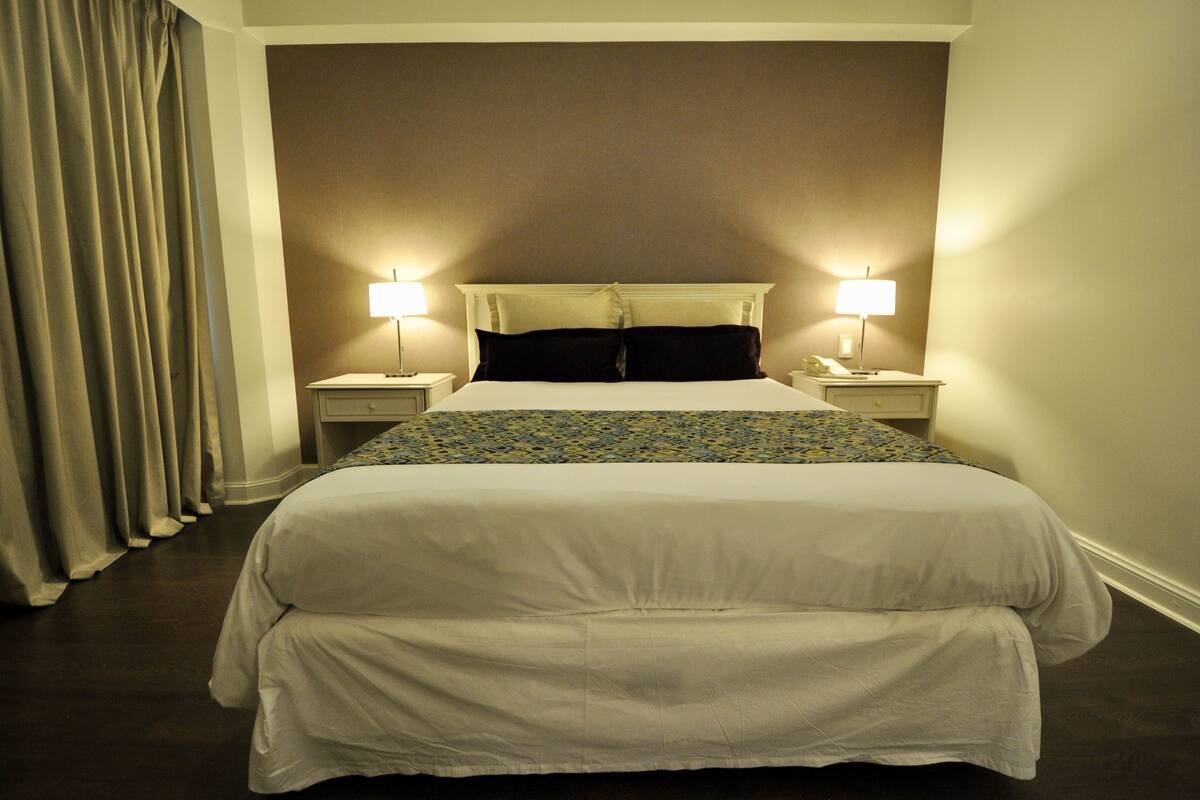 Habitació d'hotel amb un llit gran, dues làmpades de nit, un capçal de llit i una cortina.