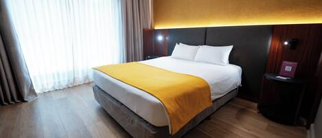 Una habitación de hotel con una cama grande, piso de madera, una manta amarilla, una mesita de noche con lámpara y una ventana con cortinas.