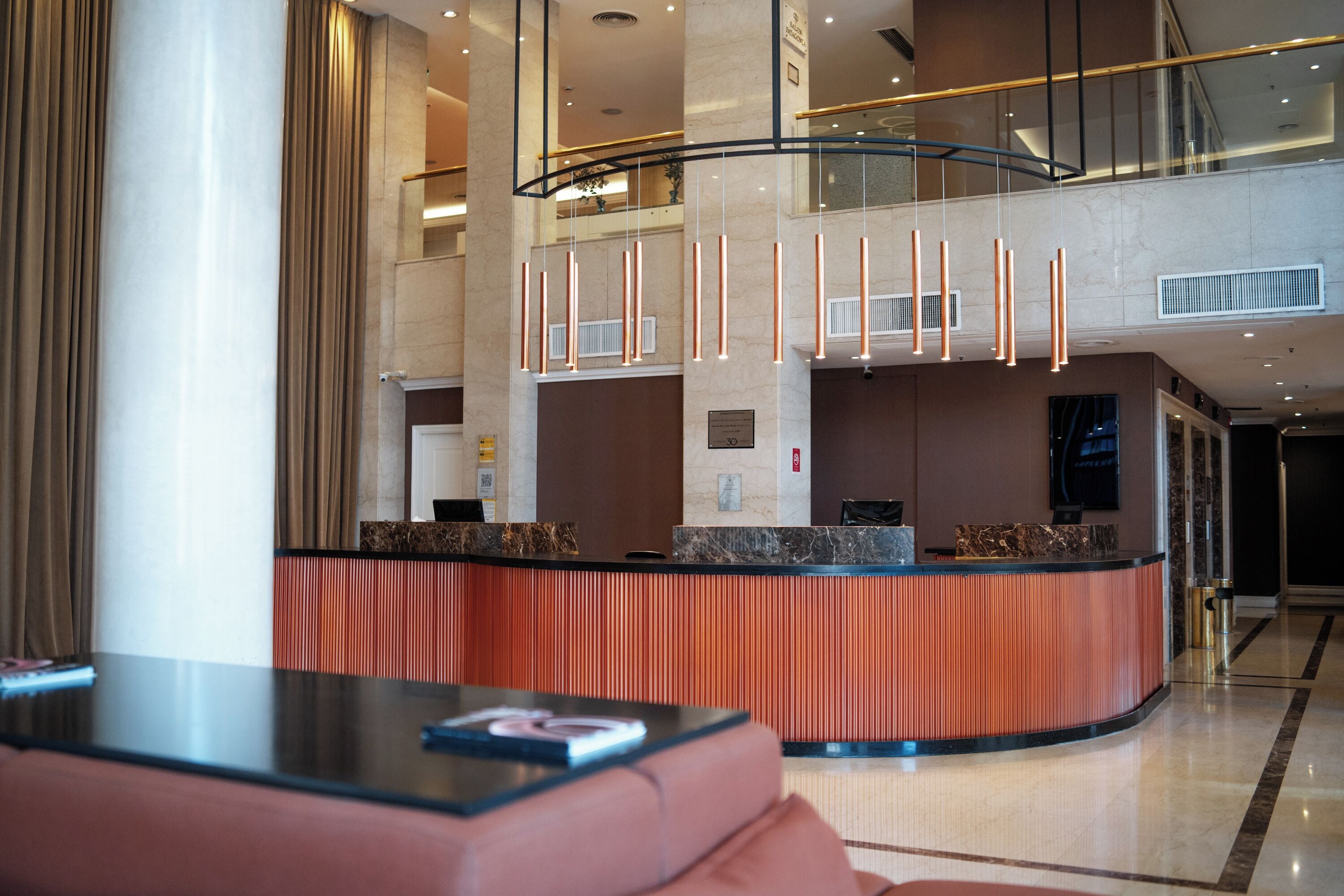 A modern hotel lobby with a curved reception desk, a seating area with a red sofa, and a high ceiling with hanging lights.