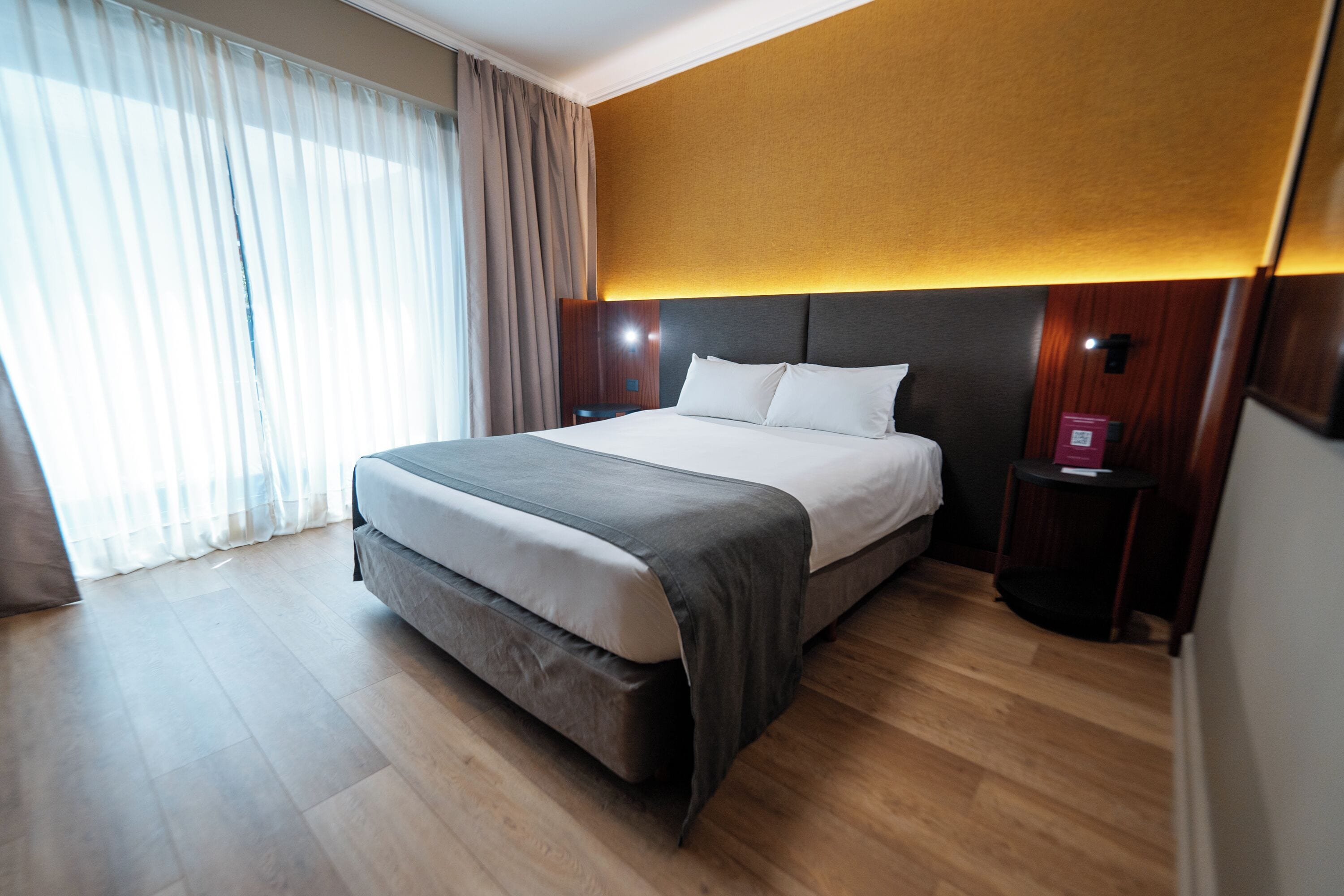 Une chambre d’hôtel moderne dotée d’un grand lit, d’un parquet en bois, d’une tête de lit avec éclairage intégré et offrant une vue sur l’extérieur à travers des rideaux transparents.