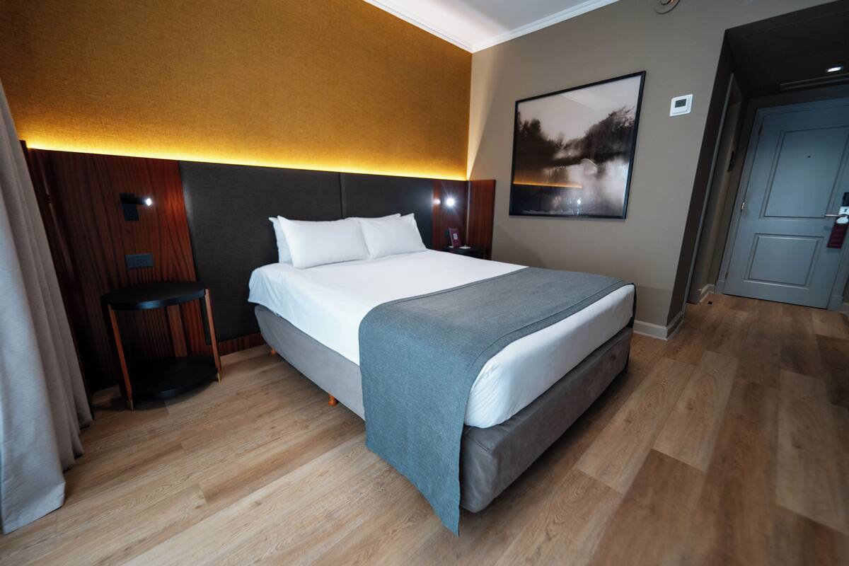 Ein Hotelzimmer mit einem großen Bett, Holzboden, einem kleinen Nachttisch, einem Beistelltisch, einer Wandleuchte und einer Tür.