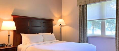 A hotel room with a bed, two bedside lamps, a wooden headboard, a window with curtains, and a patterned floor.