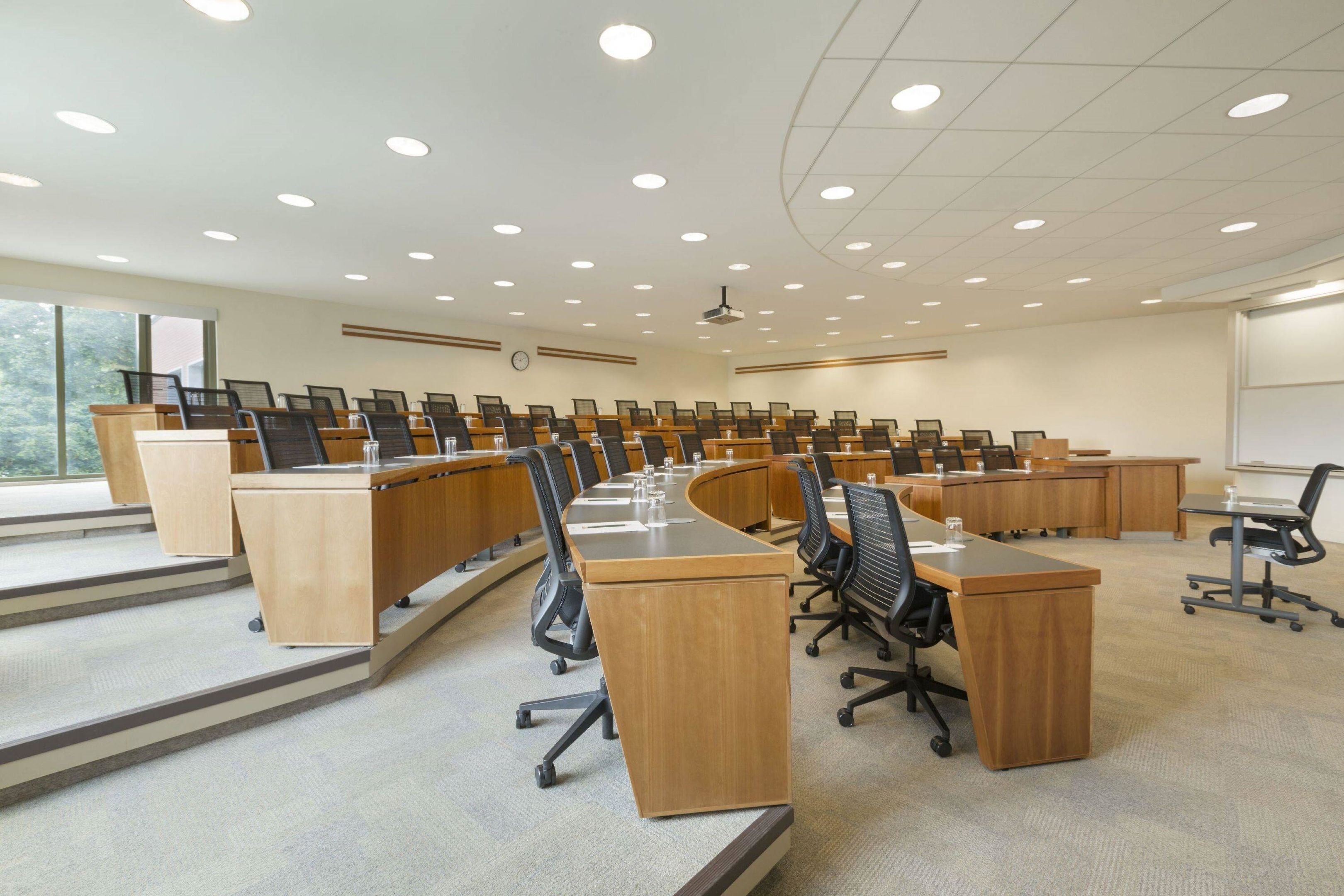 A conference room with multiple wooden desks, black office chairs, and a large screen.