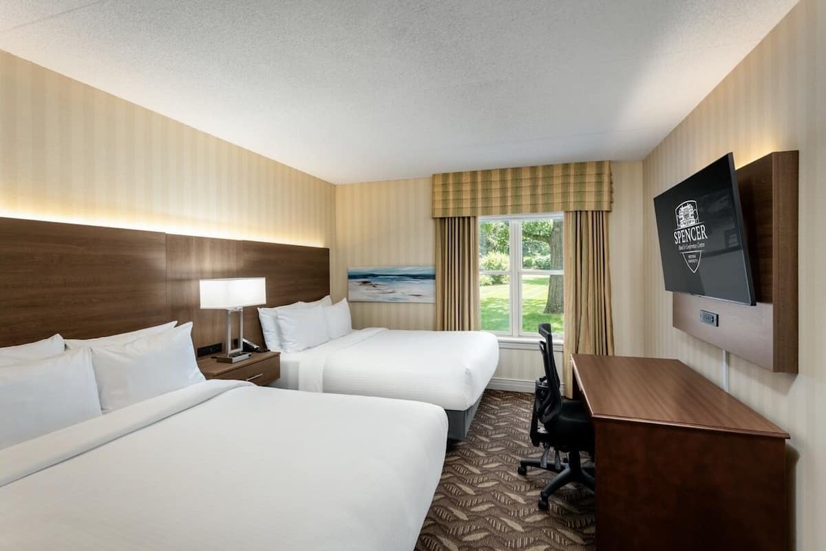 A hotel room with two beds, a desk, a TV, and a window with curtains.