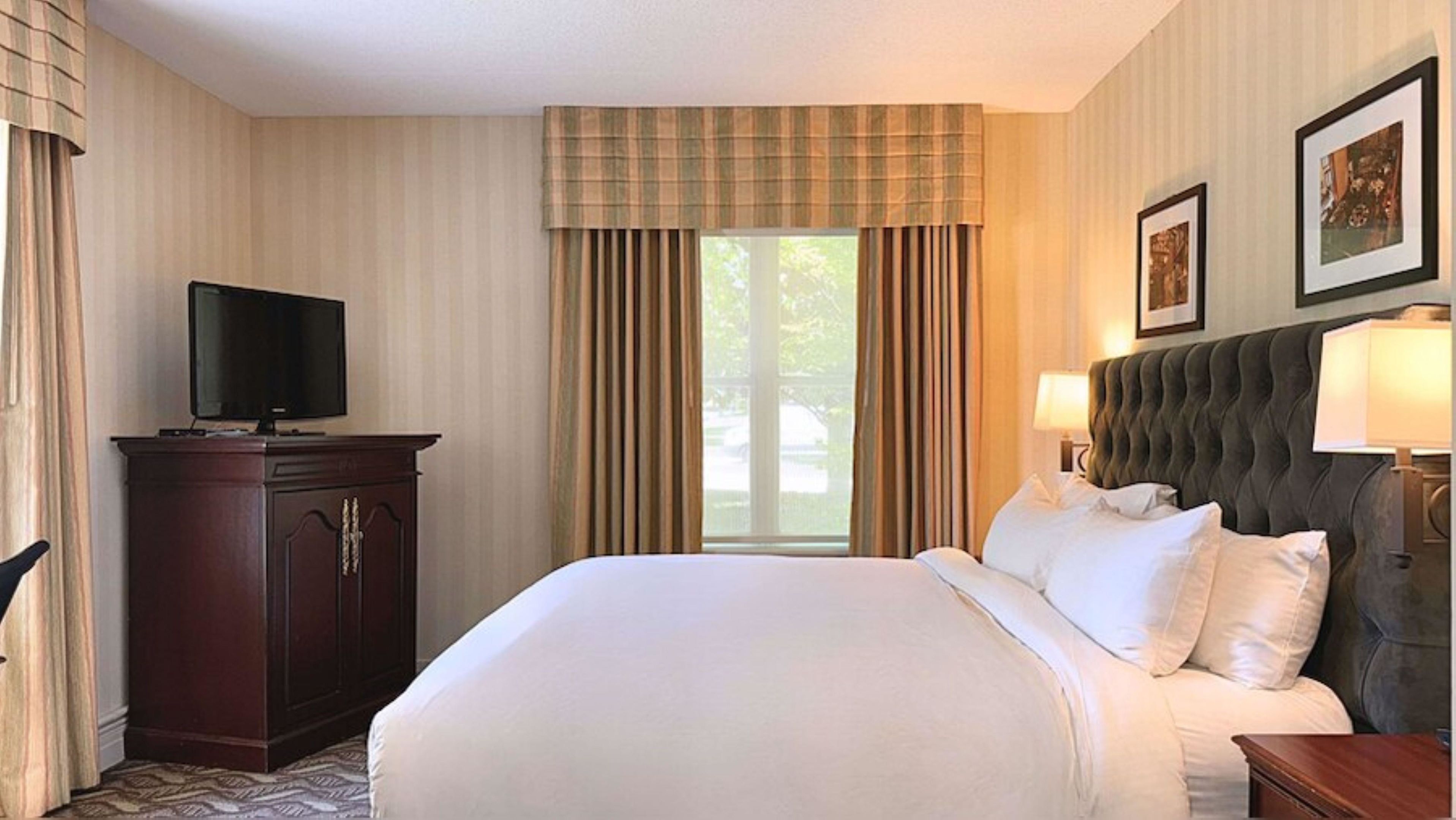 A hotel room with a bed, a television on a cabinet, a window with curtains, and a framed picture on the wall.