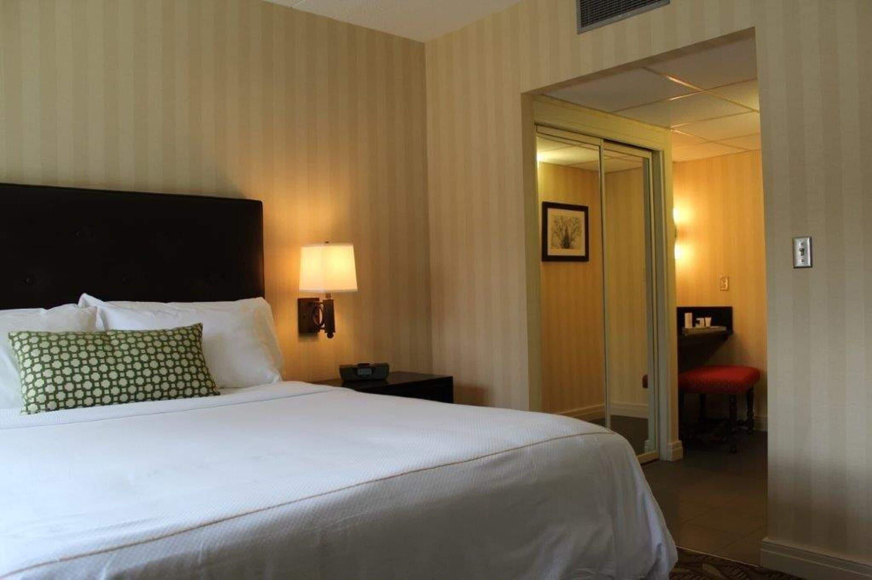 A hotel room with a large bed, a nightstand with a lamp, a red chair, and a door leading to another room.
