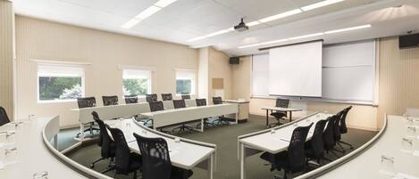 A conference room with long tables, chairs, a projector screen, and windows.