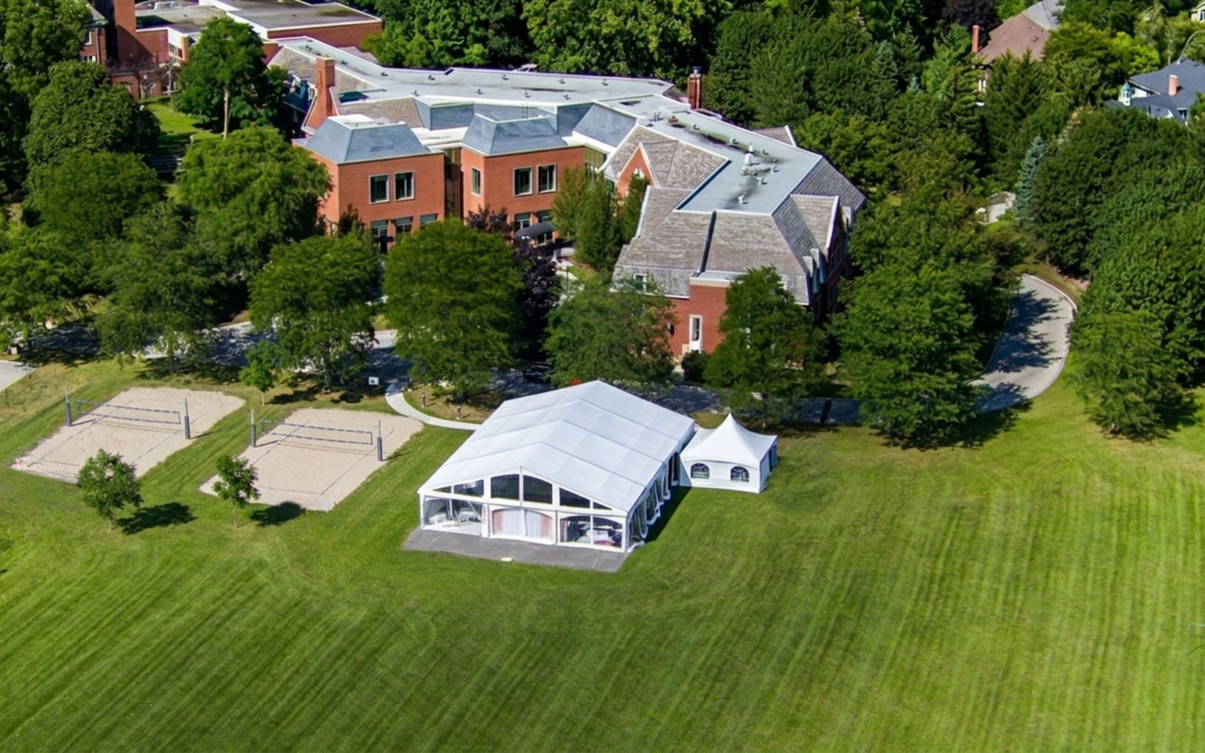 An aerial view of a residential area with a large house featuring a sloped roof, a tennis court, and a well-maintained lawn.