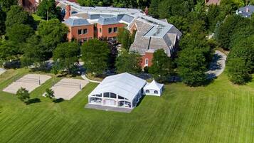 An aerial view of a residential area with a large house featuring a sloped roof, a tennis court, and a well-maintained lawn.