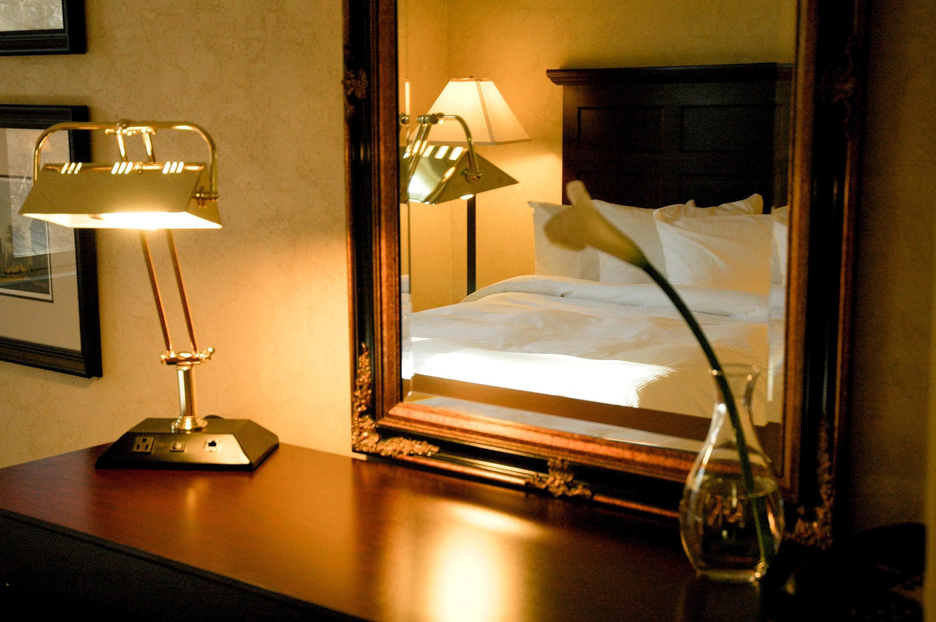 A hotel room with a bed, a nightstand with a lamp, and a mirror reflecting the room.