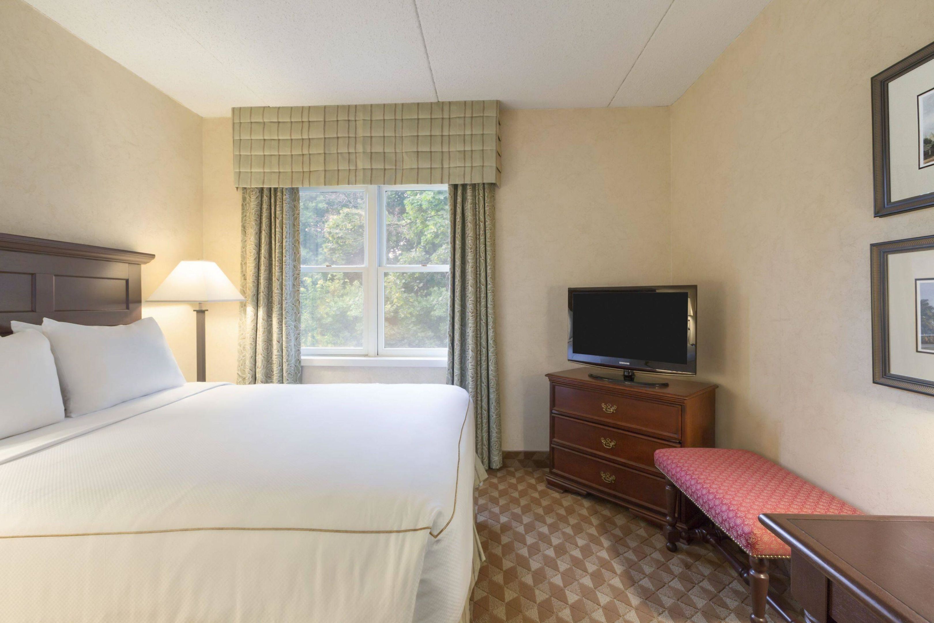 Een hotelkamer met een bed, een televisie, een dressoir, een bankje en een raam met gordijnen.