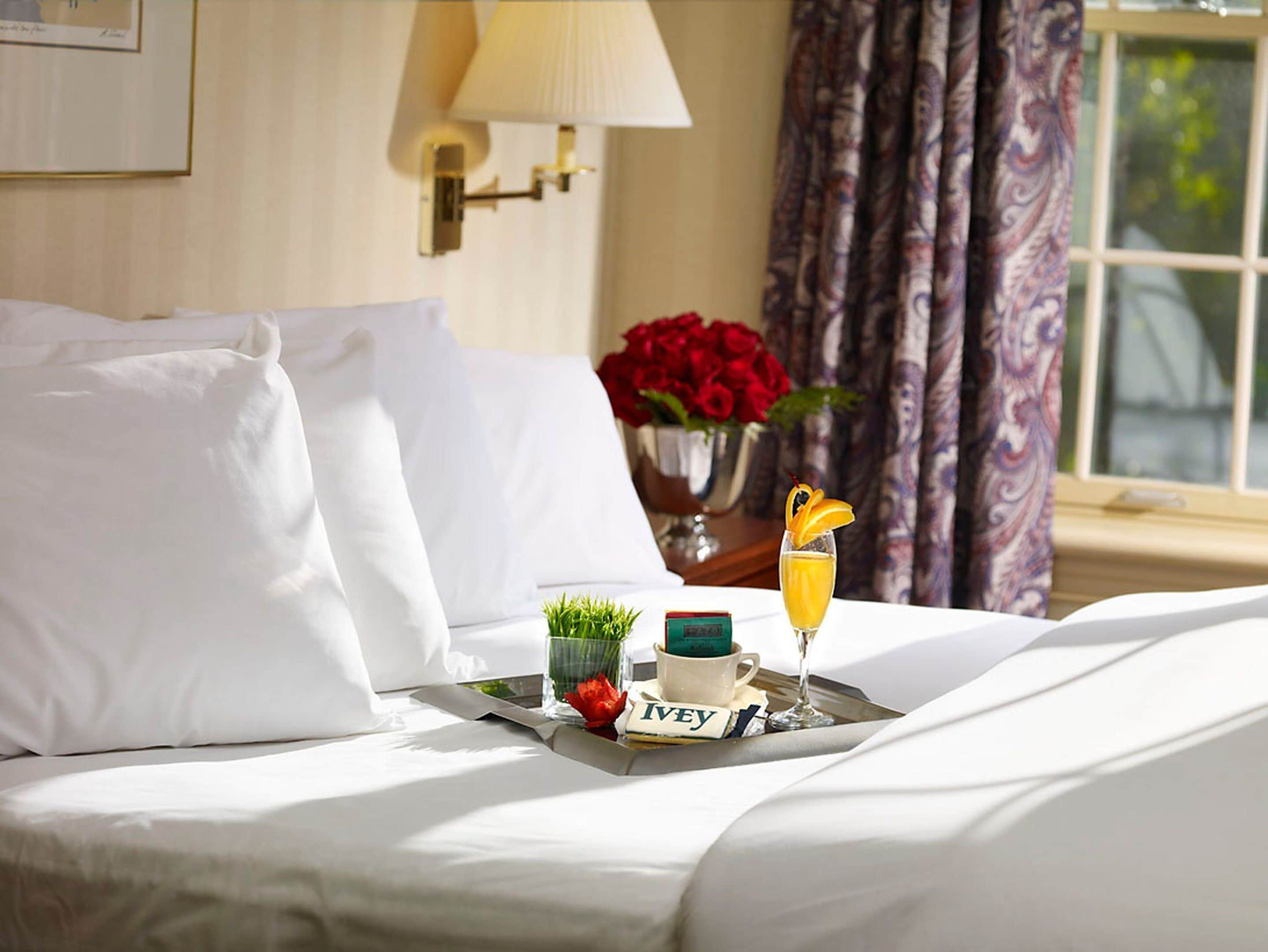 A hotel room with a neatly made bed, a tray with a drink, a vase with red flowers, and a lamp on the wall.